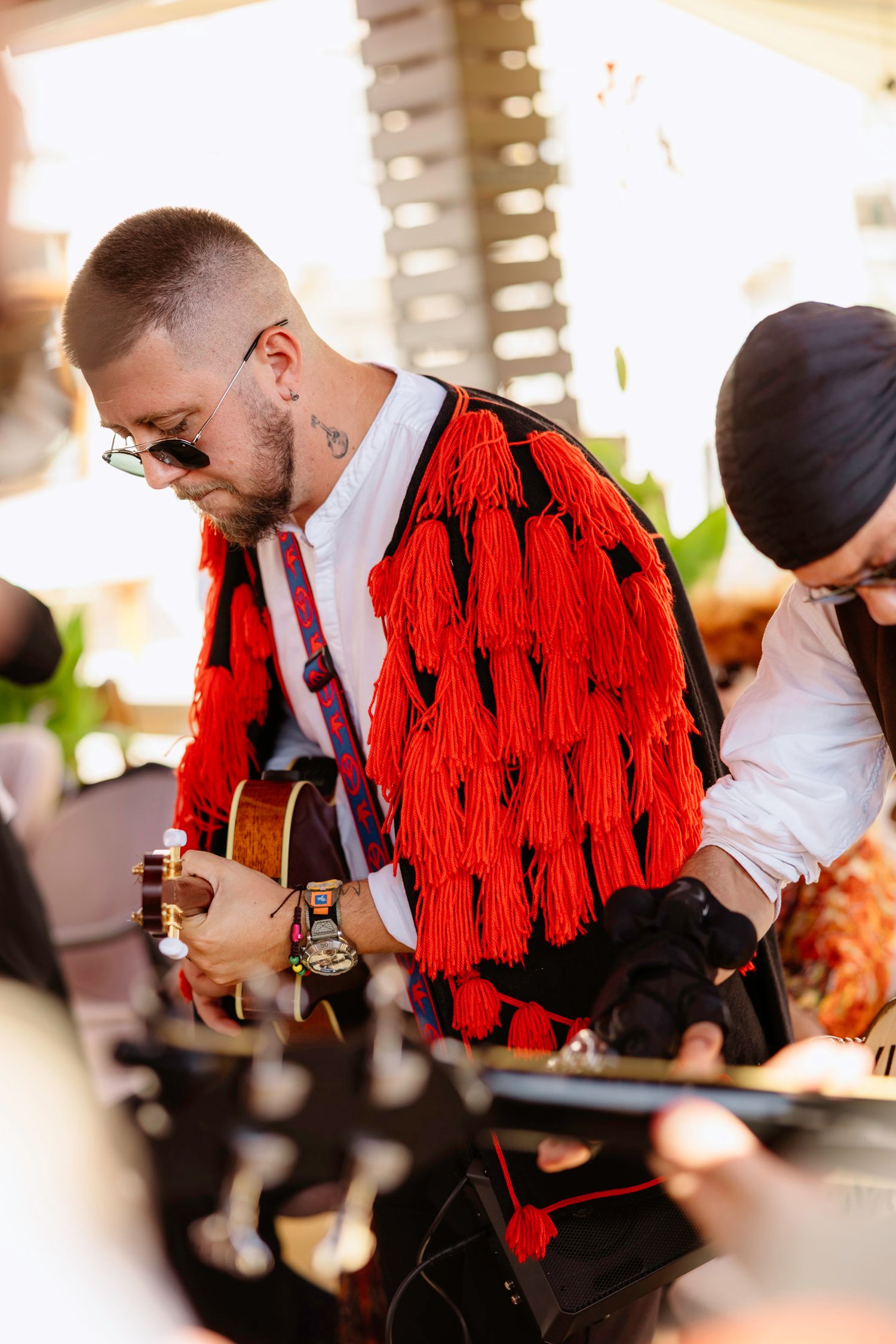 Musician wearing a striking red tasseled jacket performs with a guitar at an outdoor event.