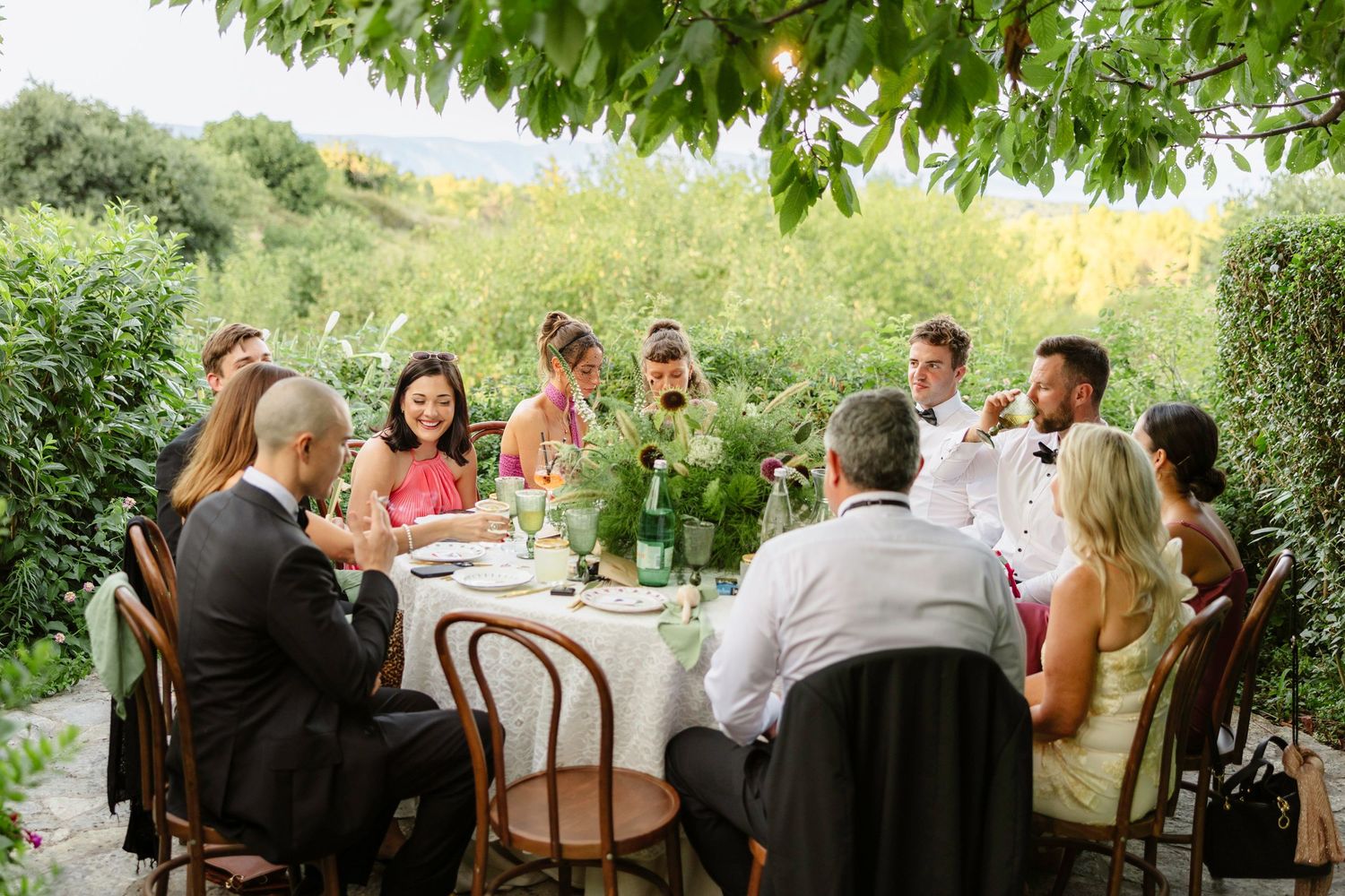 Outdoor wedding reception under green tree canopy with guests dining at long tables. Hvar, Croatia