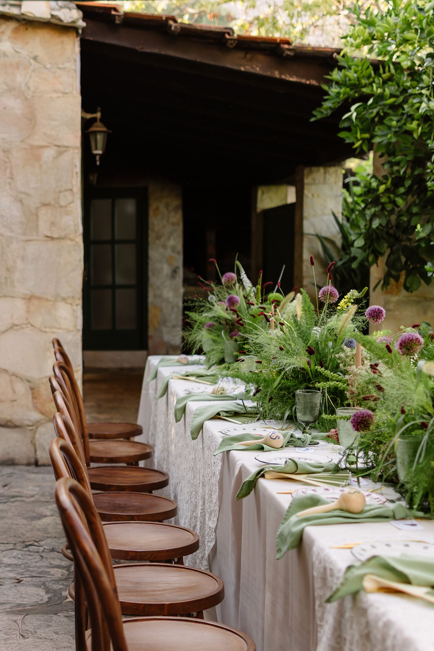 Elegant outdoor dining setup with wooden chairs and white tablecloths decorated with fresh greenery and purple flowers.