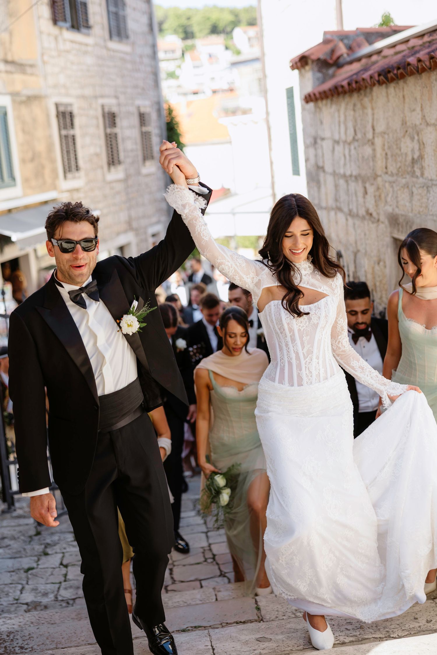 Newlyweds walking hand in hand down cobblestone streets with wedding party following behind.