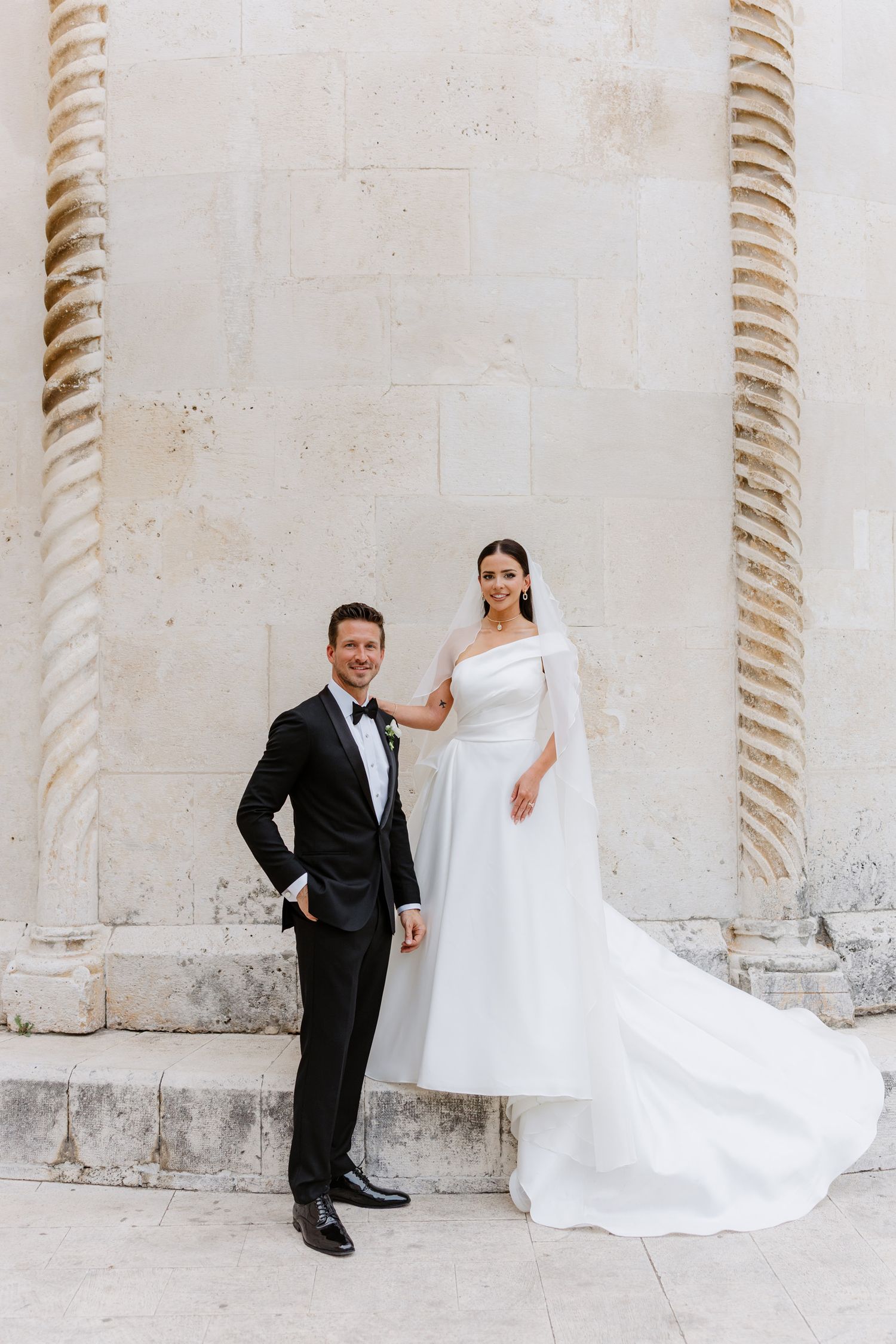 Bride in flowing white dress and groom in black suit pose playfully between ornate white stone columns.