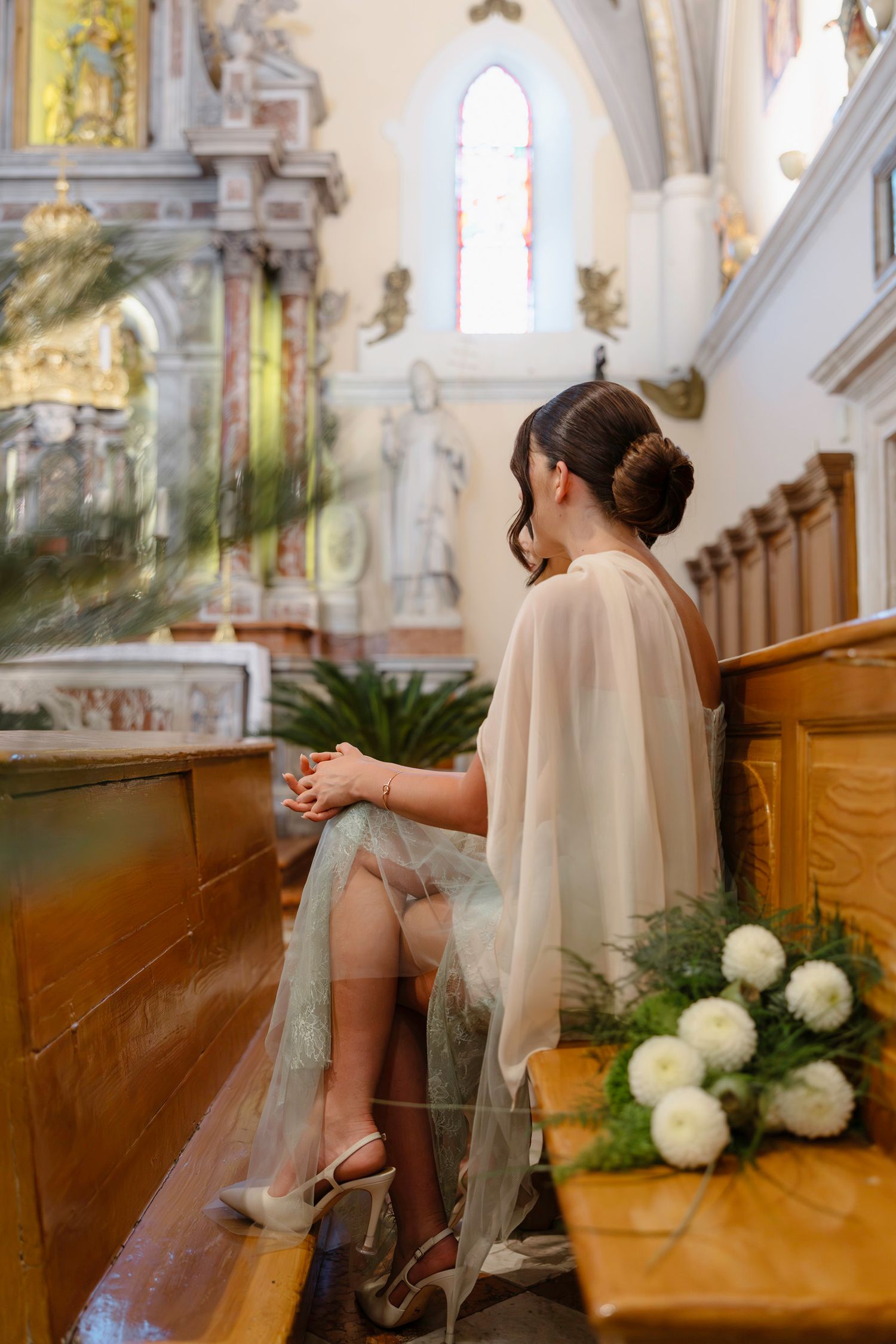 Bride sitting in wooden church pew with elegant flowing veil and white floral decorations.