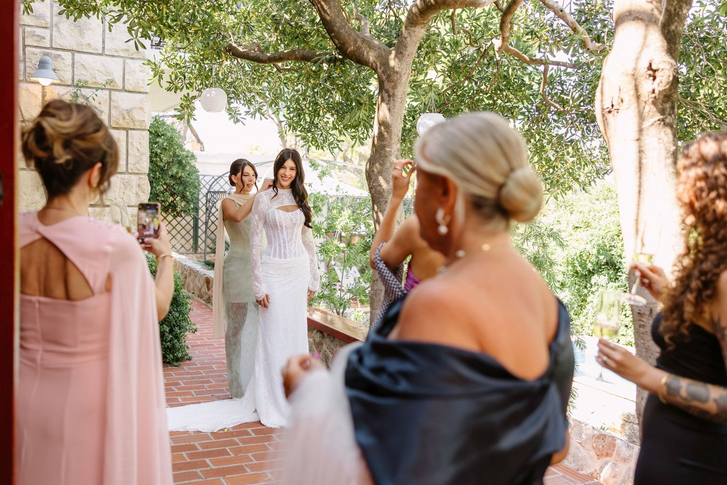 Wedding party members gather under a tree-covered courtyard with brick walkways.