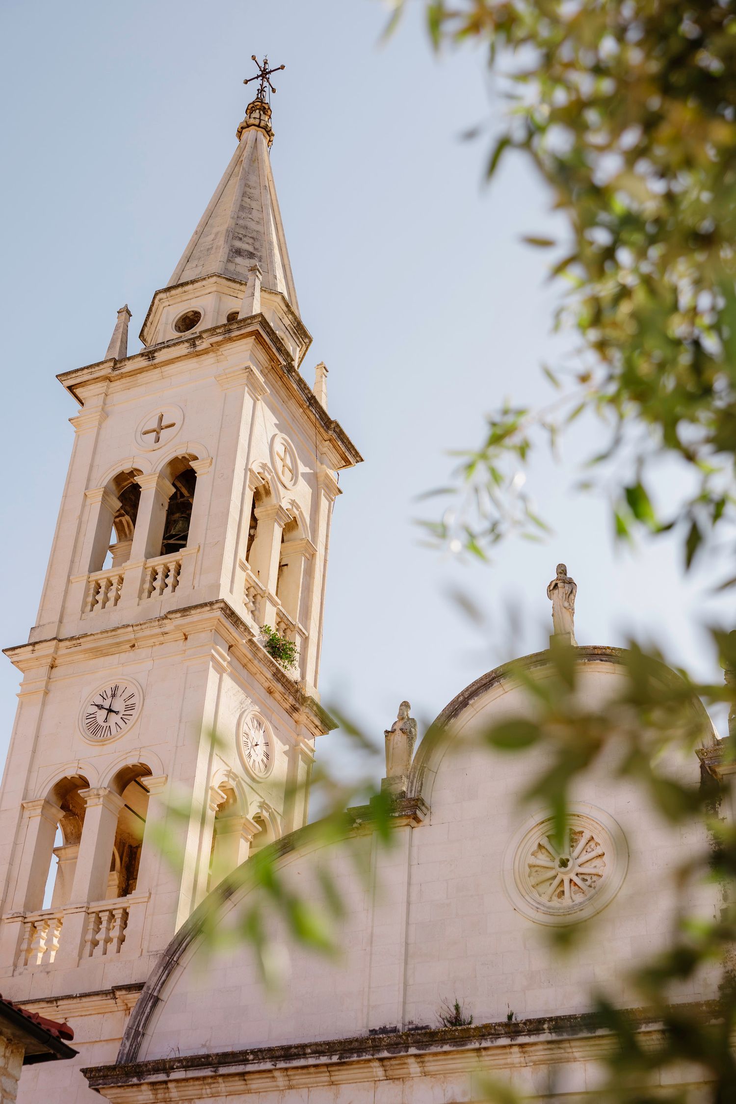 White church bell tower with clock and cross against blue sky, framed by olive tree branches.