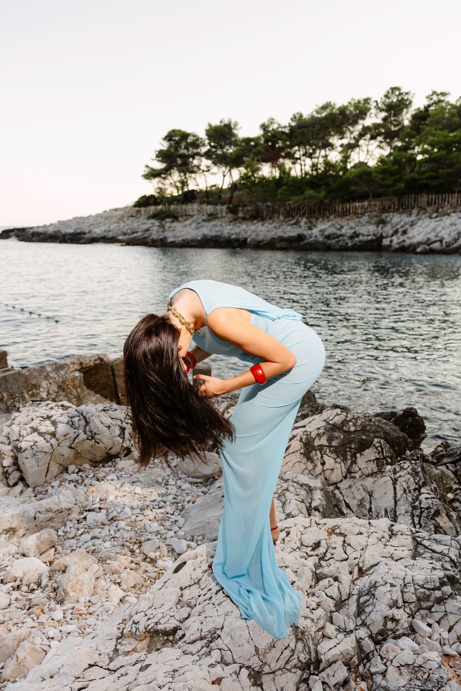 A woman in a flowing blue dress dances playfully on coastal rocks by the water.