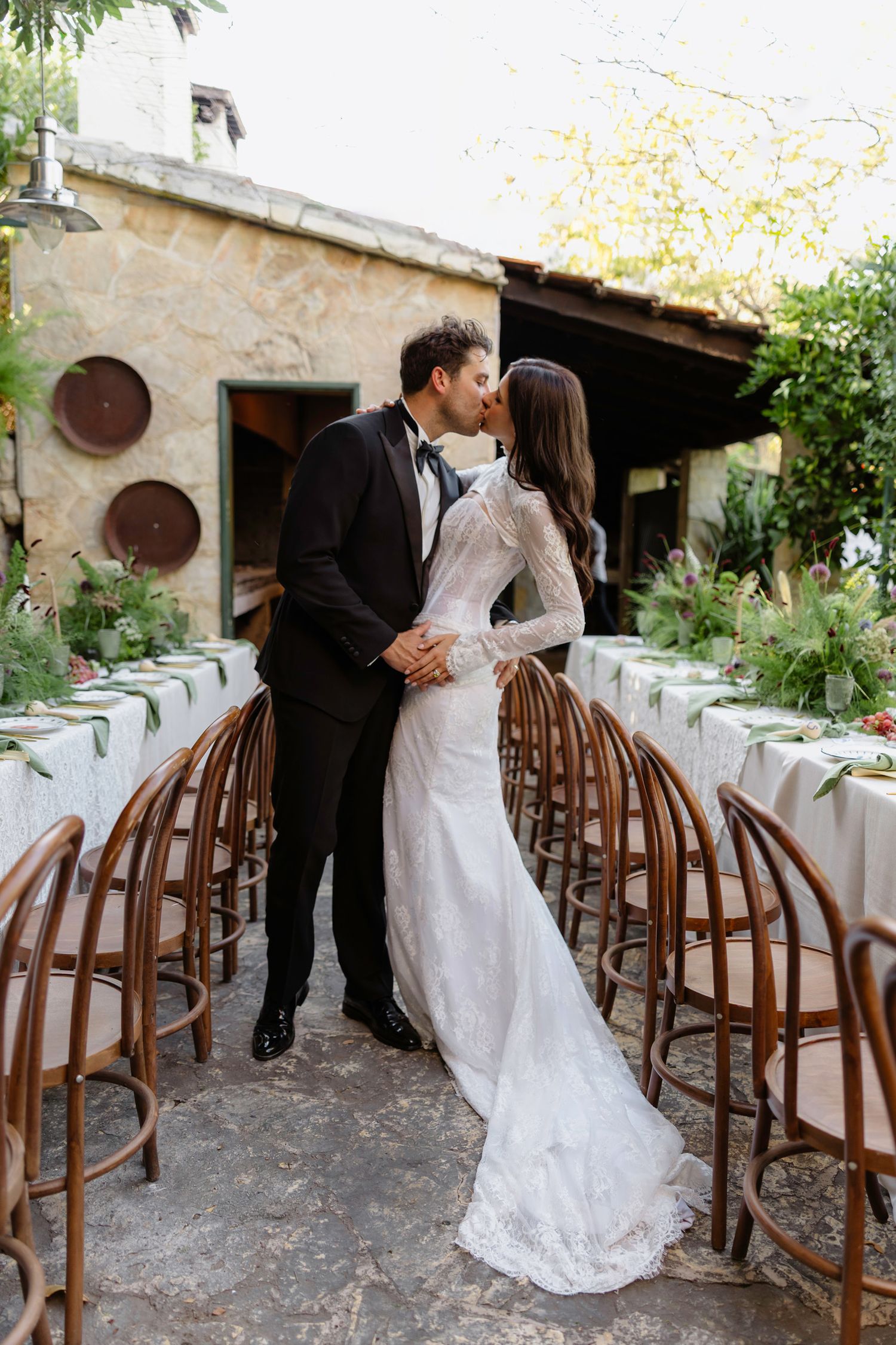 Wedding couple embracing by rustic stone wall with elegant table settings and wooden chairs.