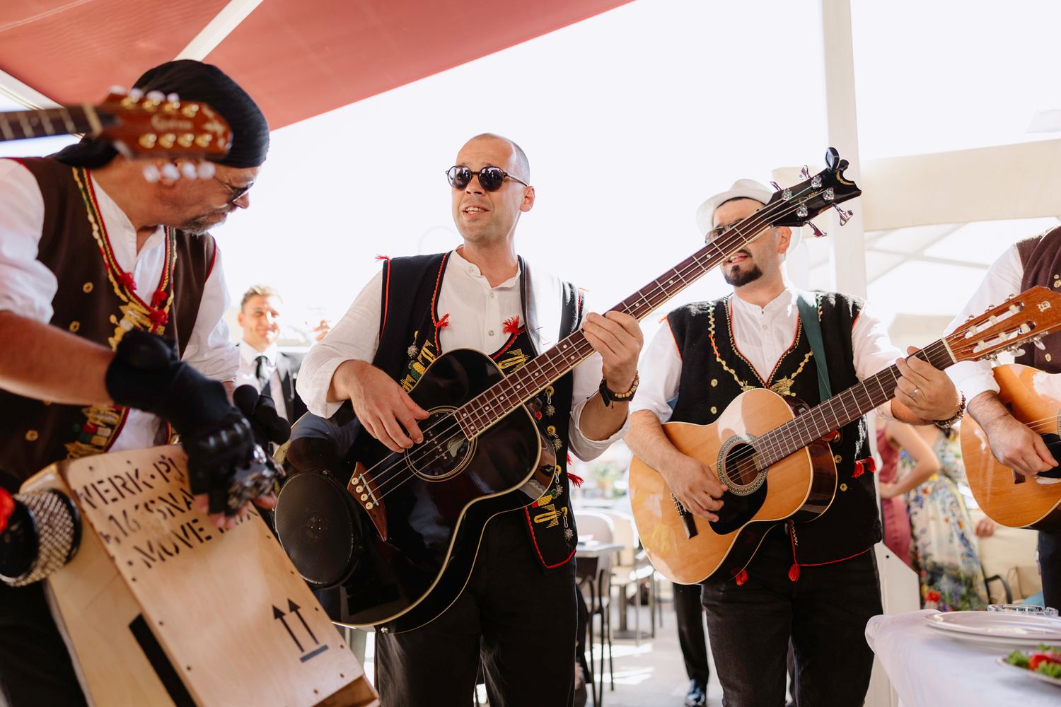 Band members perform with guitars at an outdoor wedding reception under white canopies.
