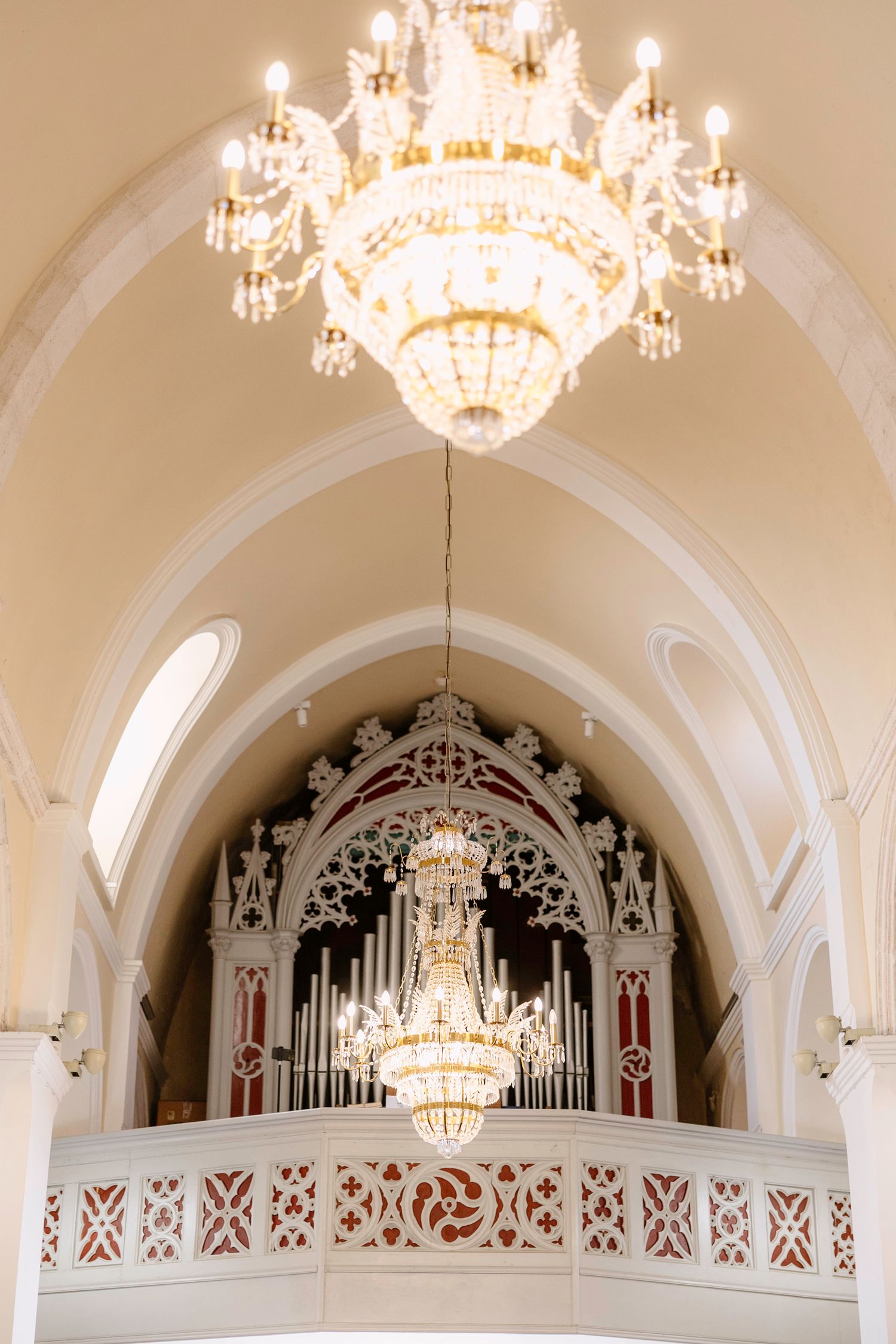 Grand crystal chandeliers hanging from ornate white arched ceiling with decorative organ pipes.