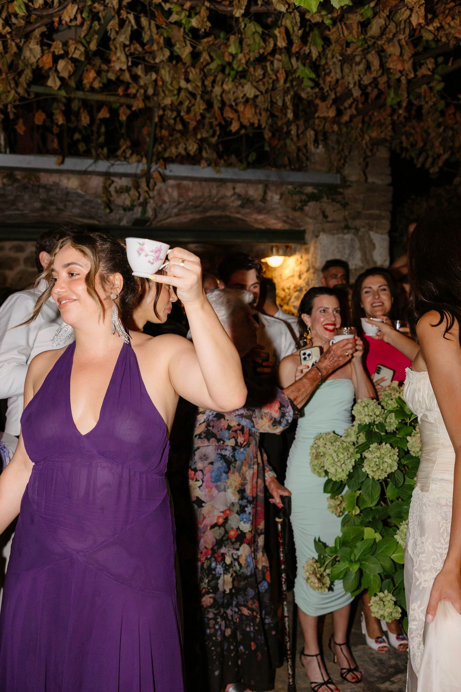 Wedding guests in purple and light colored dresses dancing together at an evening celebration.
