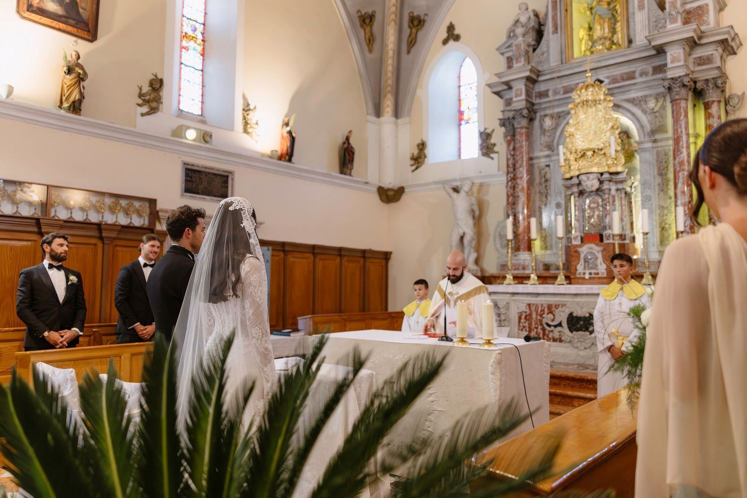 Interior of an ornate church during a wedding ceremony with priest at altar and decorative palm plants.