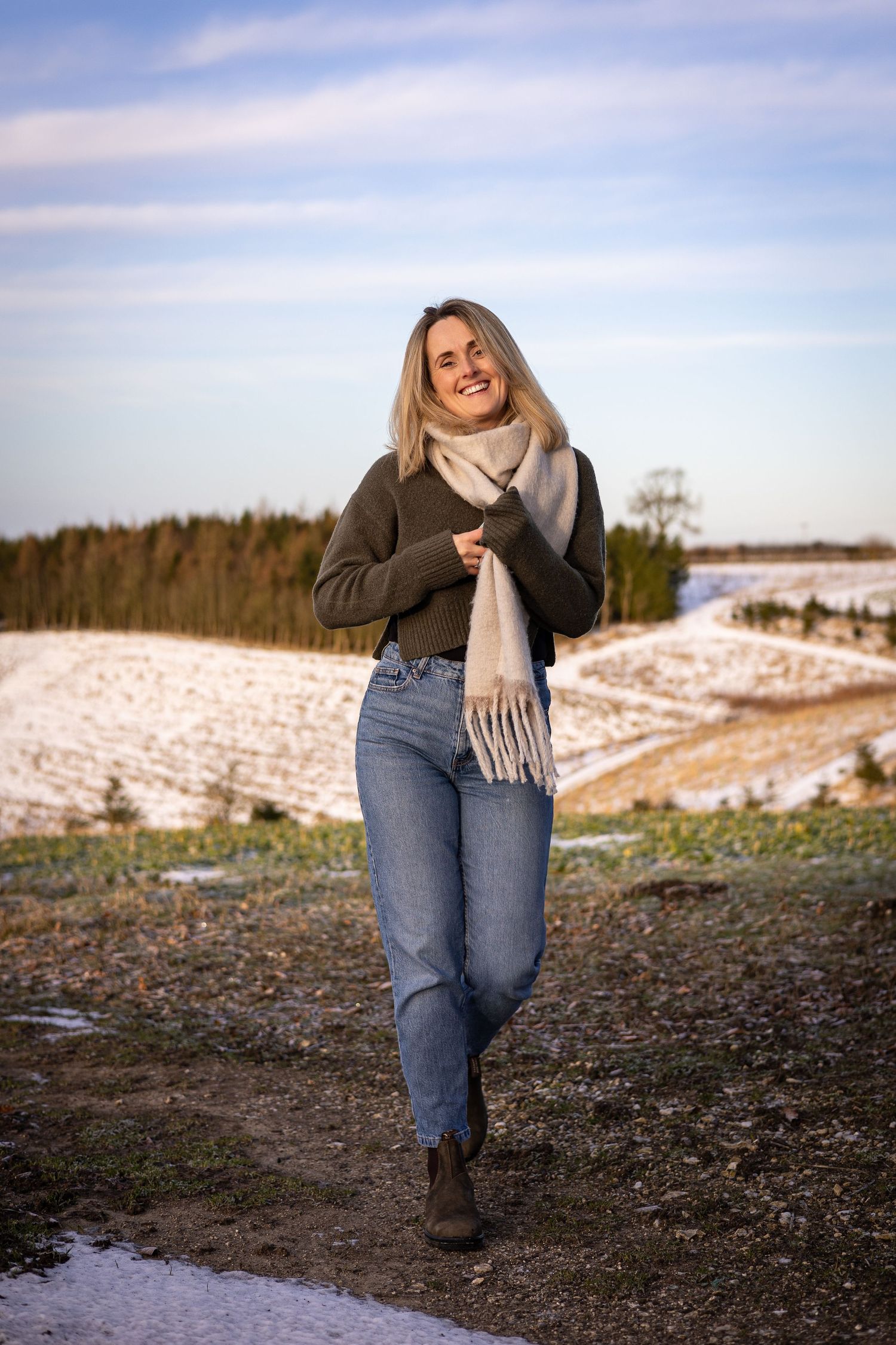 A winter outdoor portrait of someone in a cozy sweater, scarf and jeans against a snowy landscape.