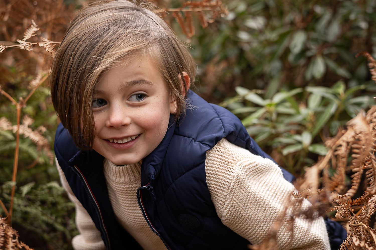 Child in navy jacket and cream sweater smiling among autumn foliage.