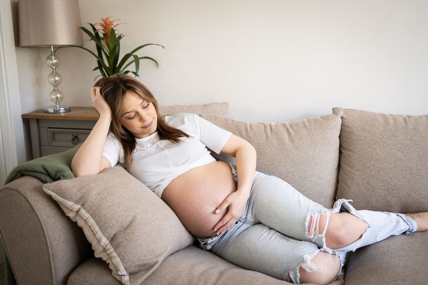 Pregnant person relaxing on a beige couch wearing ripped jeans and a white shirt with a houseplant in the background.