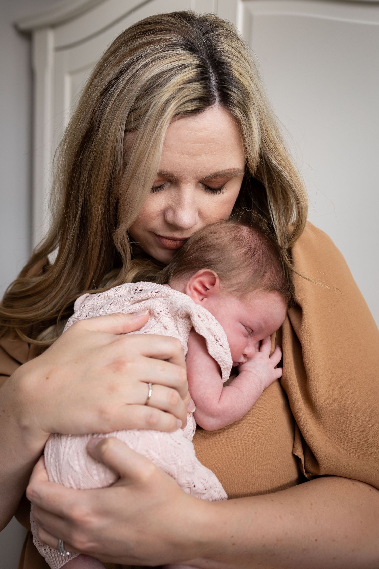 A parent wearing a tan sweater tenderly embraces and holds a sleeping newborn baby wearing a pink lace outfit.