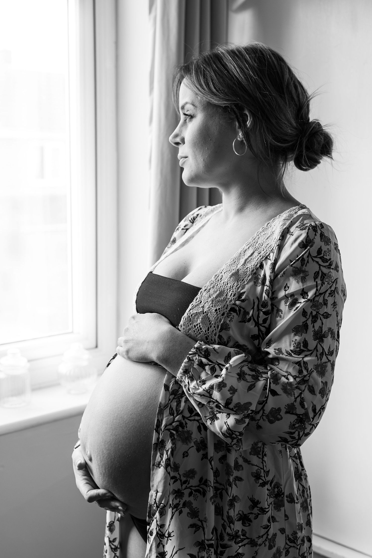 Black and white portrait of an expectant mother in a floral dress gazing thoughtfully out a window.