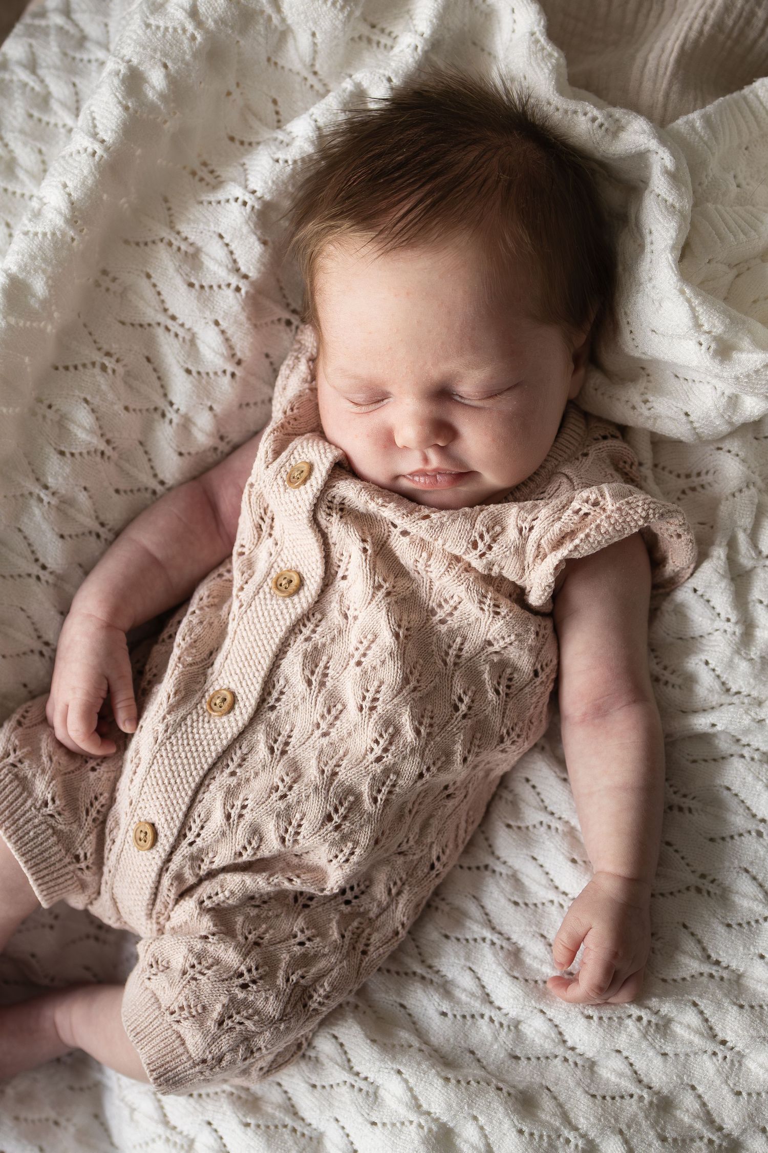 A sleeping baby lies peacefully on white lace bedding wearing a soft knit romper with wood buttons.