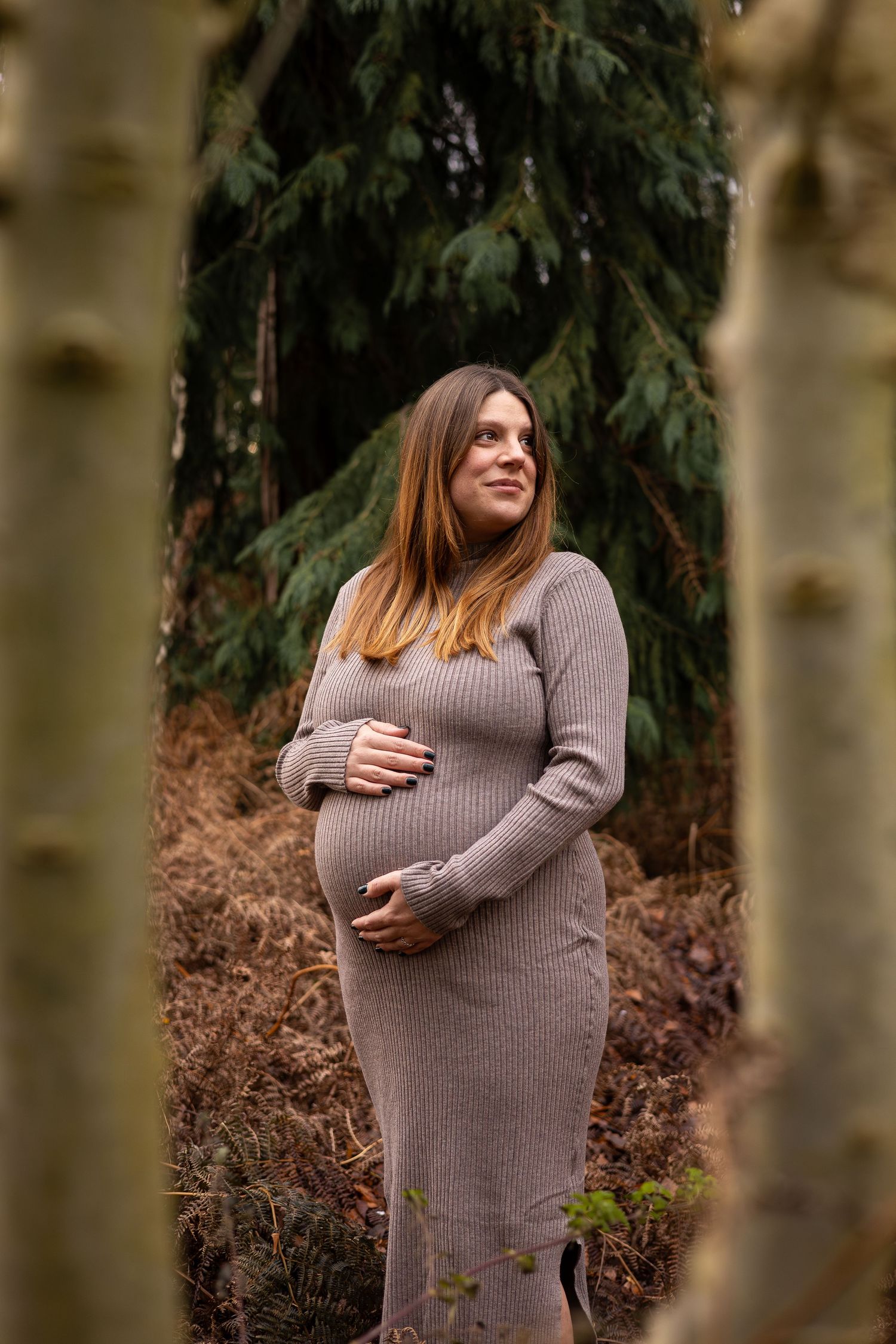 Expectant mother in gray sweater dress poses thoughtfully in a forest setting during maternity photoshoot.