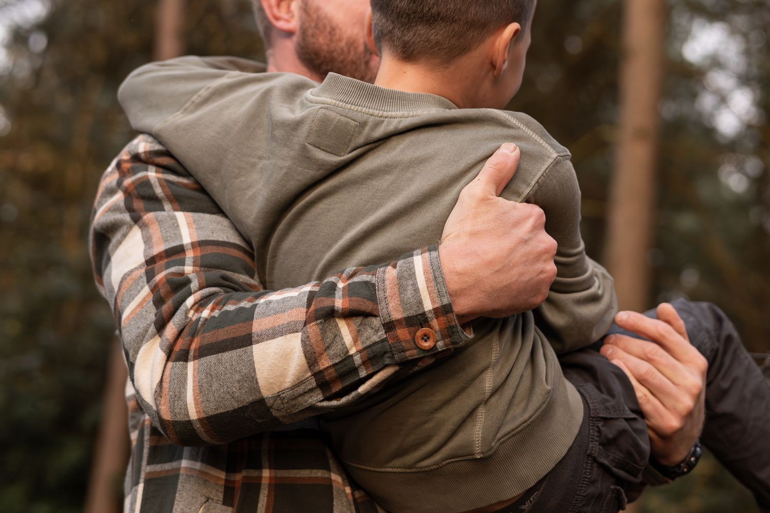A warm embrace between two people wearing plaid flannel and a green hoodie against a blurred forest background.