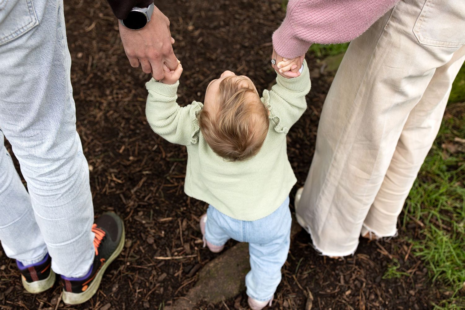 Small child learning to walk while holding hands with adults on mulch ground.