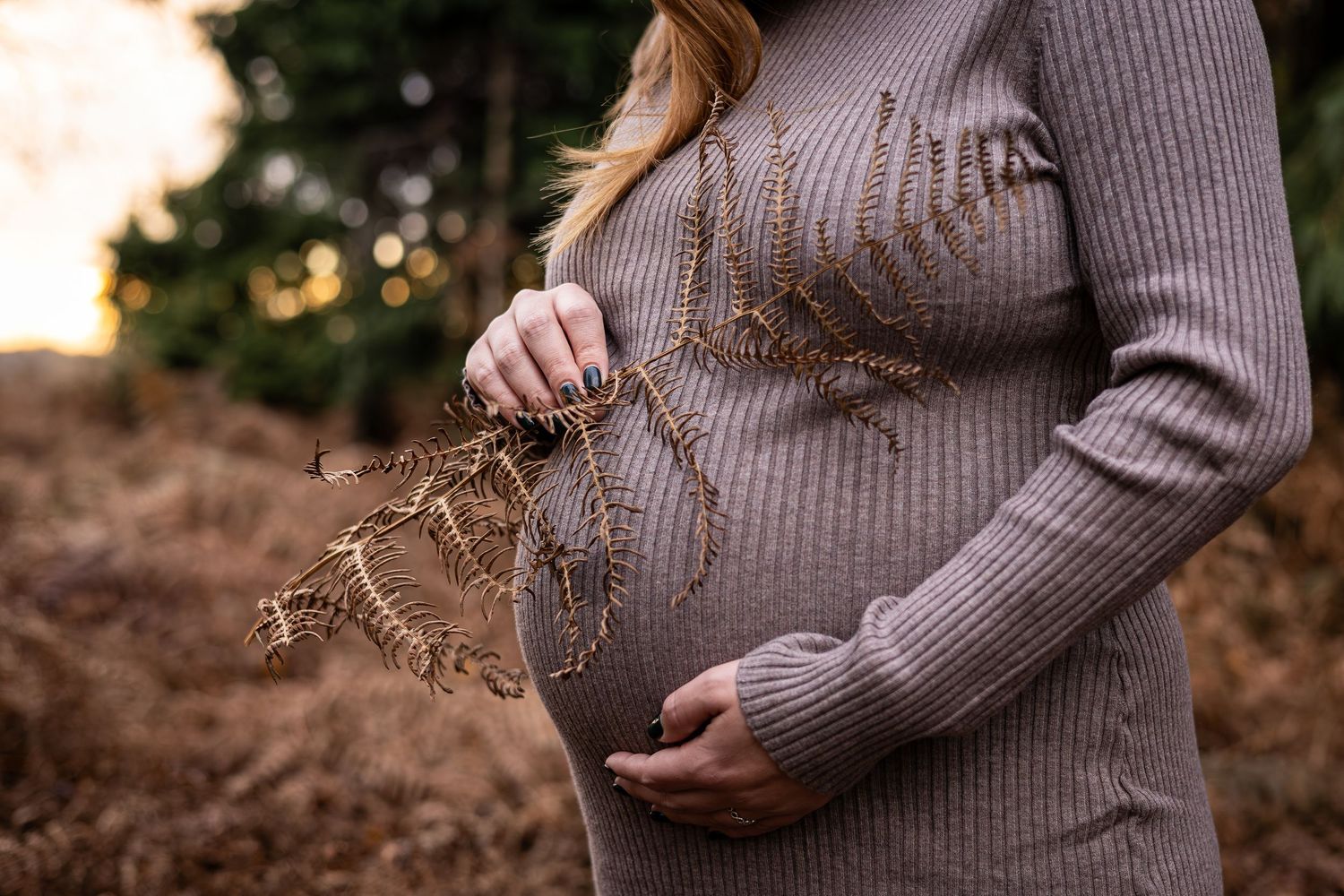 Expectant person in a brown ribbed sweater holds dried plants at sunset in a rustic outdoor setting.