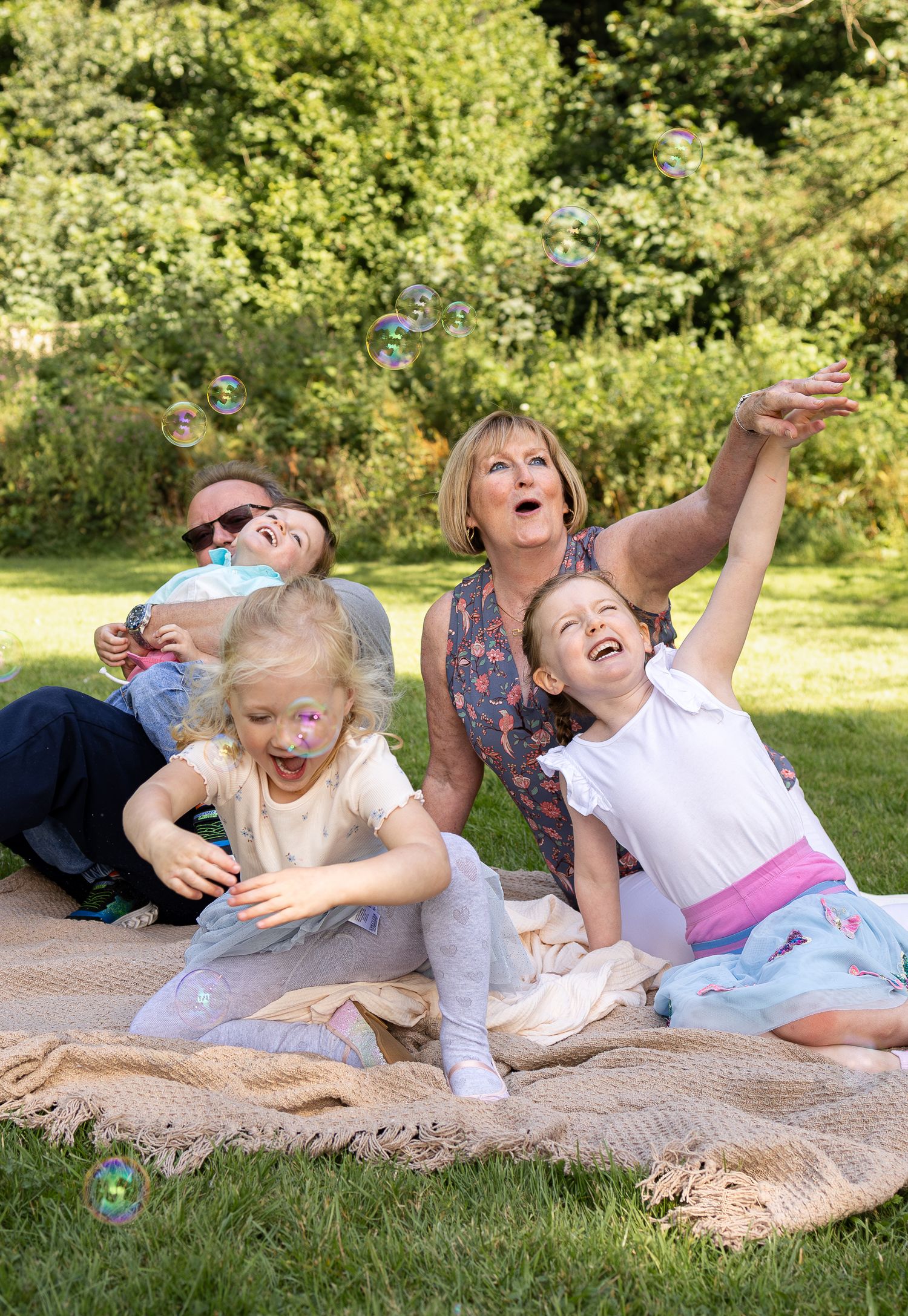 Family lying on blanket outdoors playing with bubbles and laughing together.