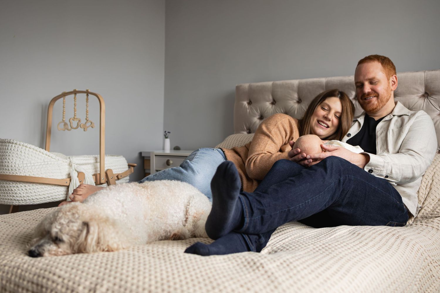 Couple relaxing on a bed with their dog while leaning against a tufted headboard in a cozy bedroom.
