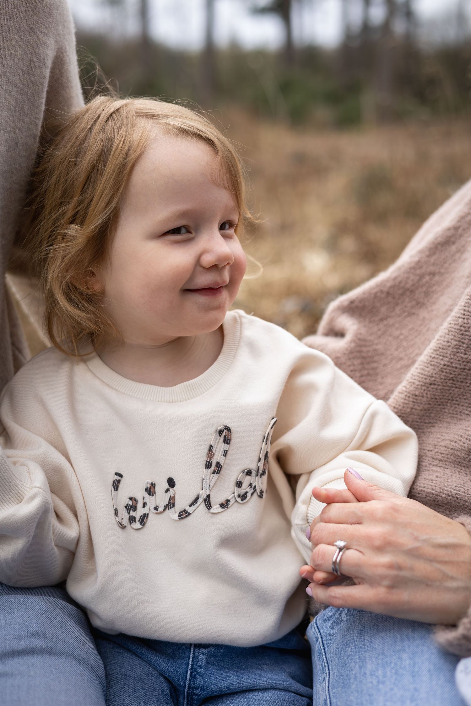 Young child in a white sweater and jeans sits happily beside an adult, enjoying a tender family moment outdoors.