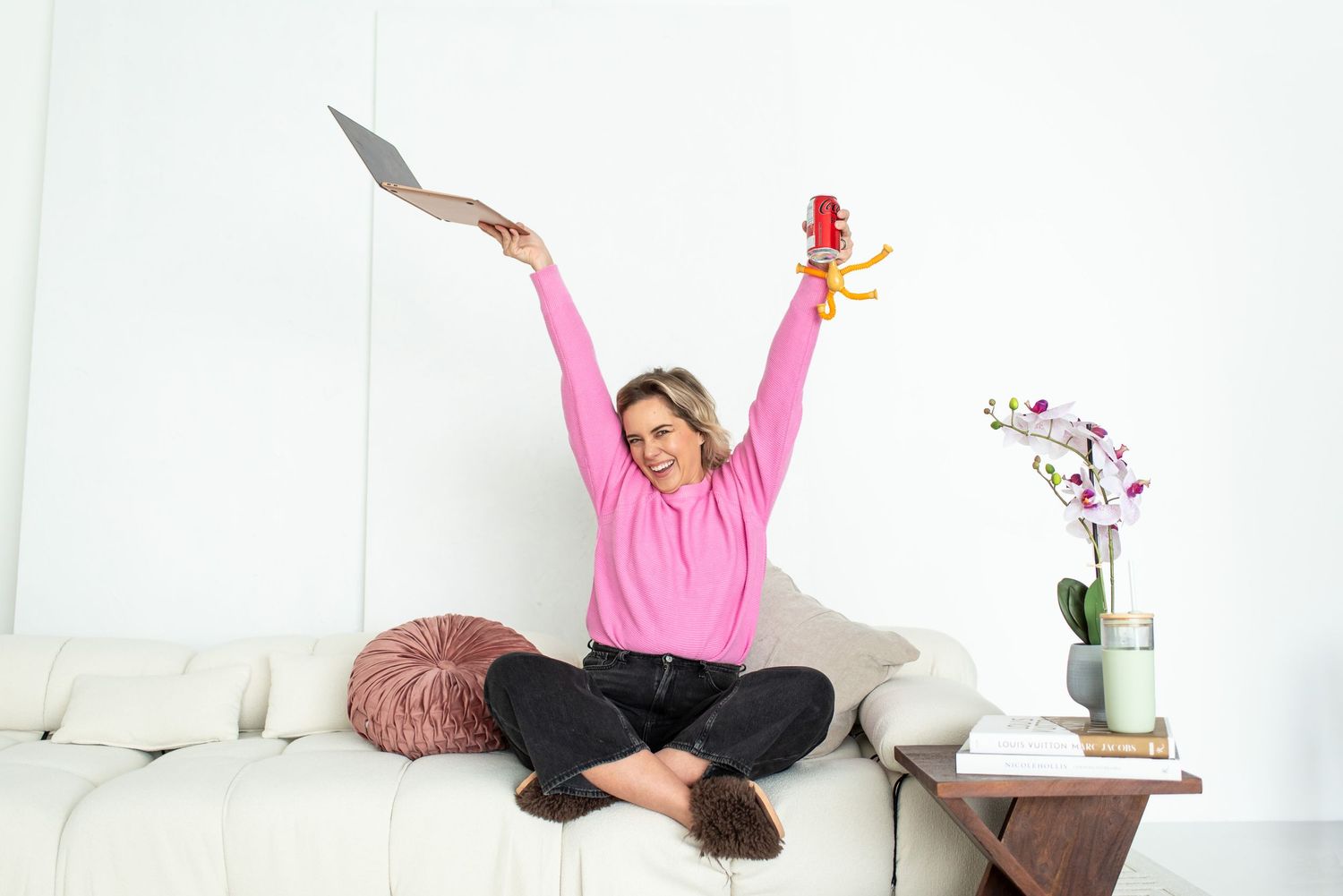 Someone wearing a pink sweater sits cross-legged on a white sofa with arms raised in celebration.