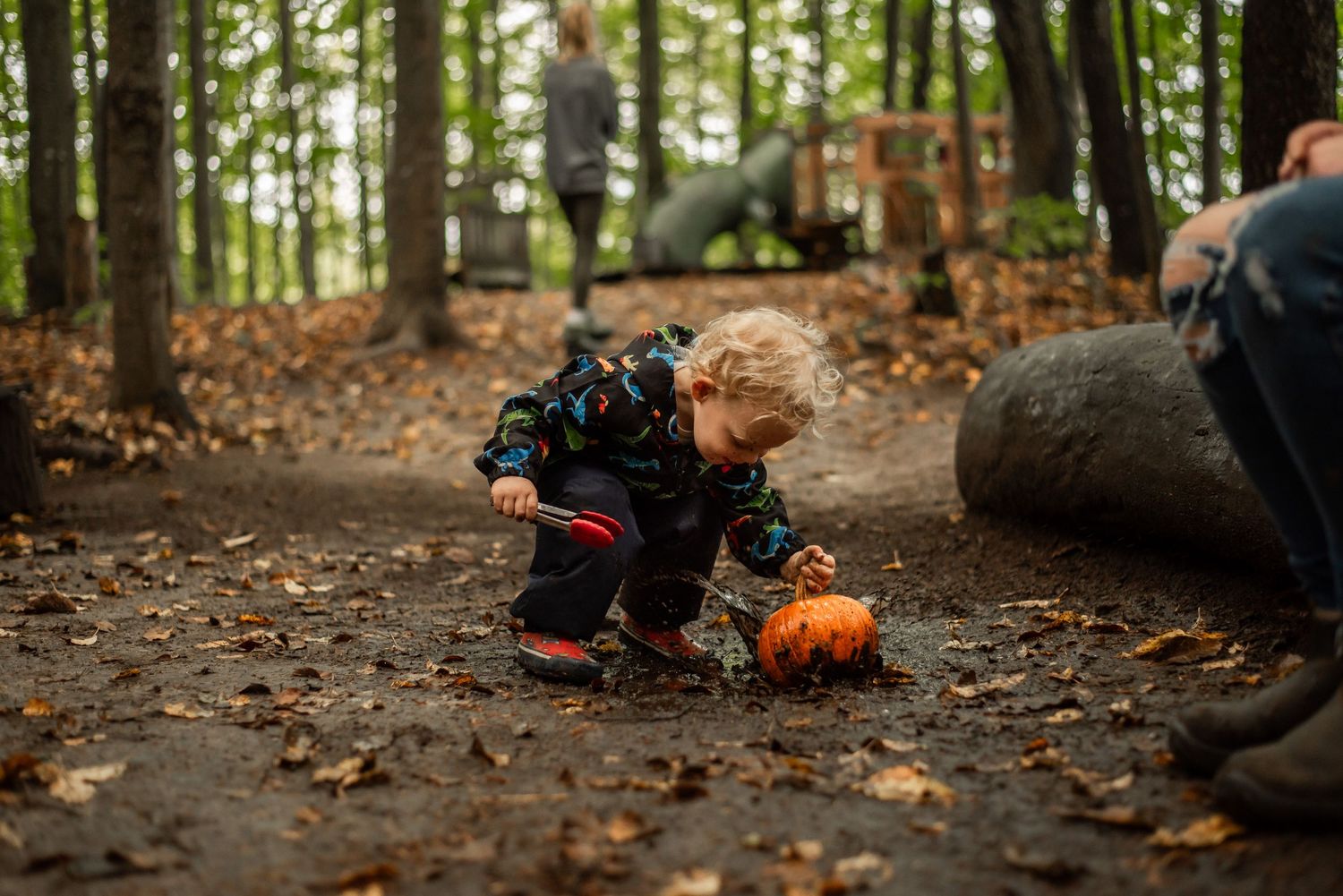 A small figure in a dragon costume crouches beside a carved pumpkin on a forest trail covered in autumn leaves.