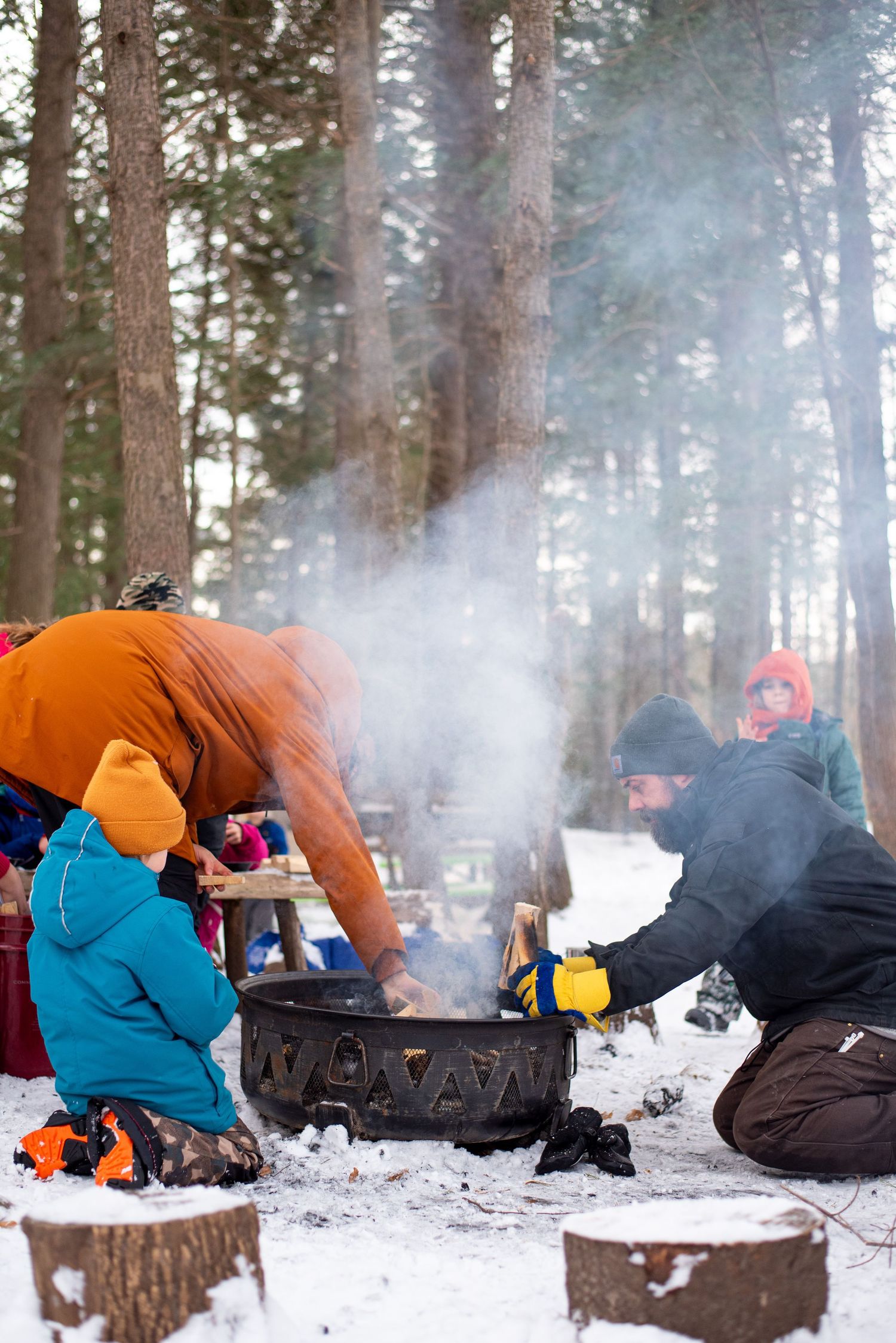 Winter campers huddle around a steaming camp stove in snowy forest while preparing a meal outdoors.