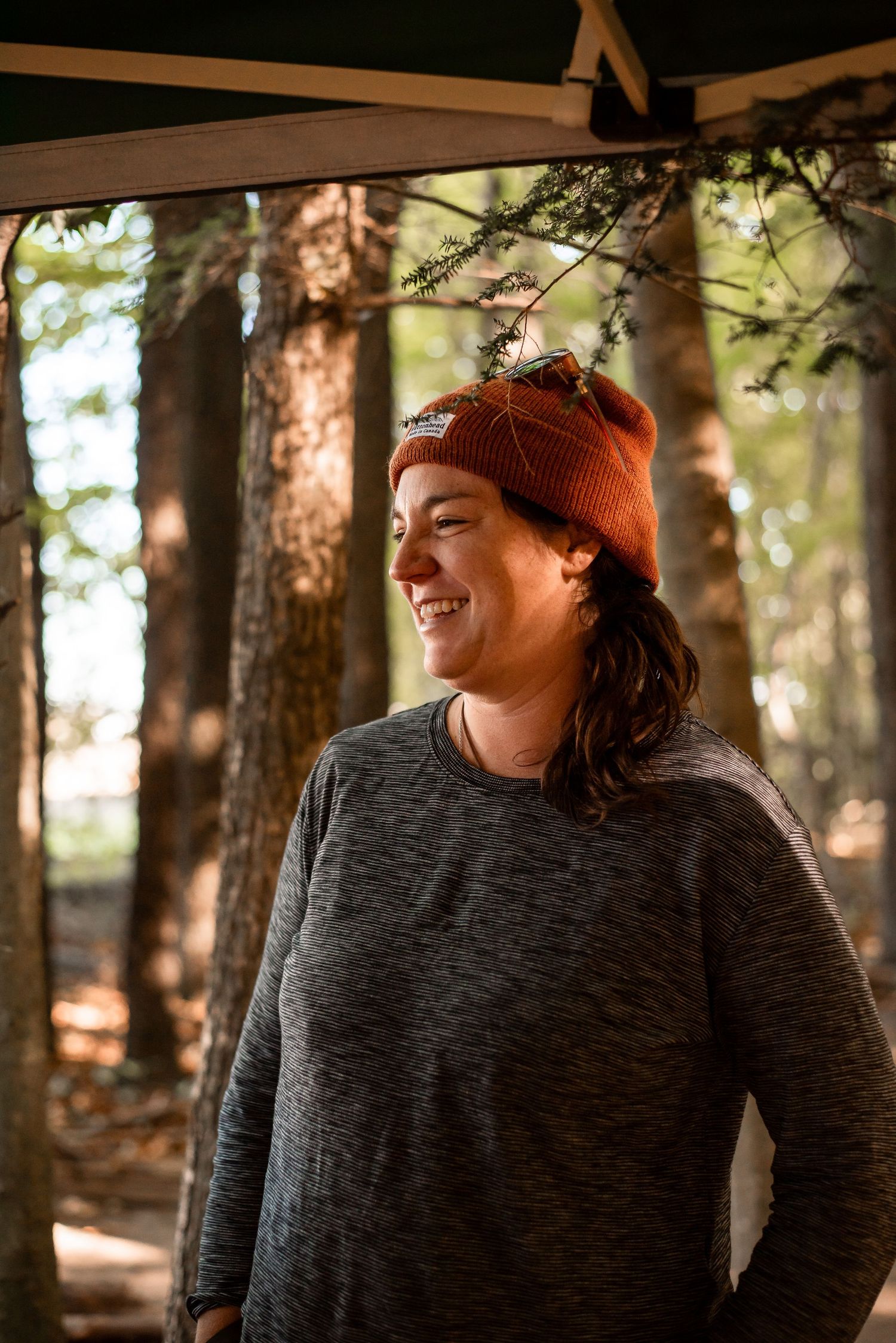 Someone wearing an orange beanie and gray sweater smiles while standing in a sunlit forest setting.