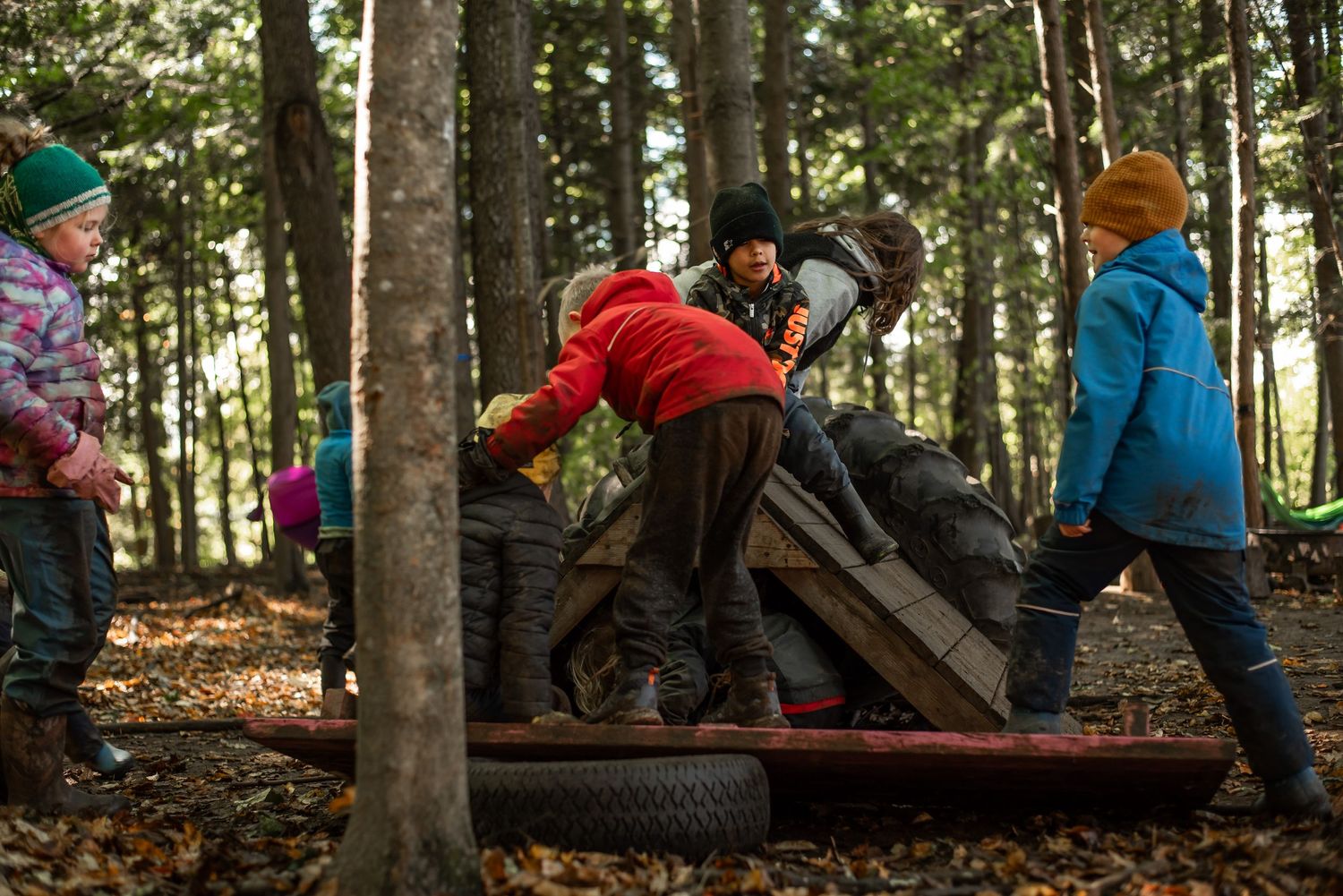 Children play on wooden logs and tires at an outdoor forest playground in autumn.