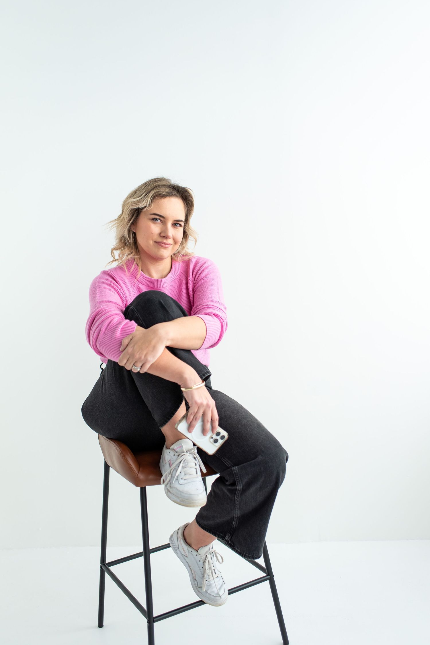 Person wearing a pink sweater and dark pants sits casually on a black bar stool against a white background.