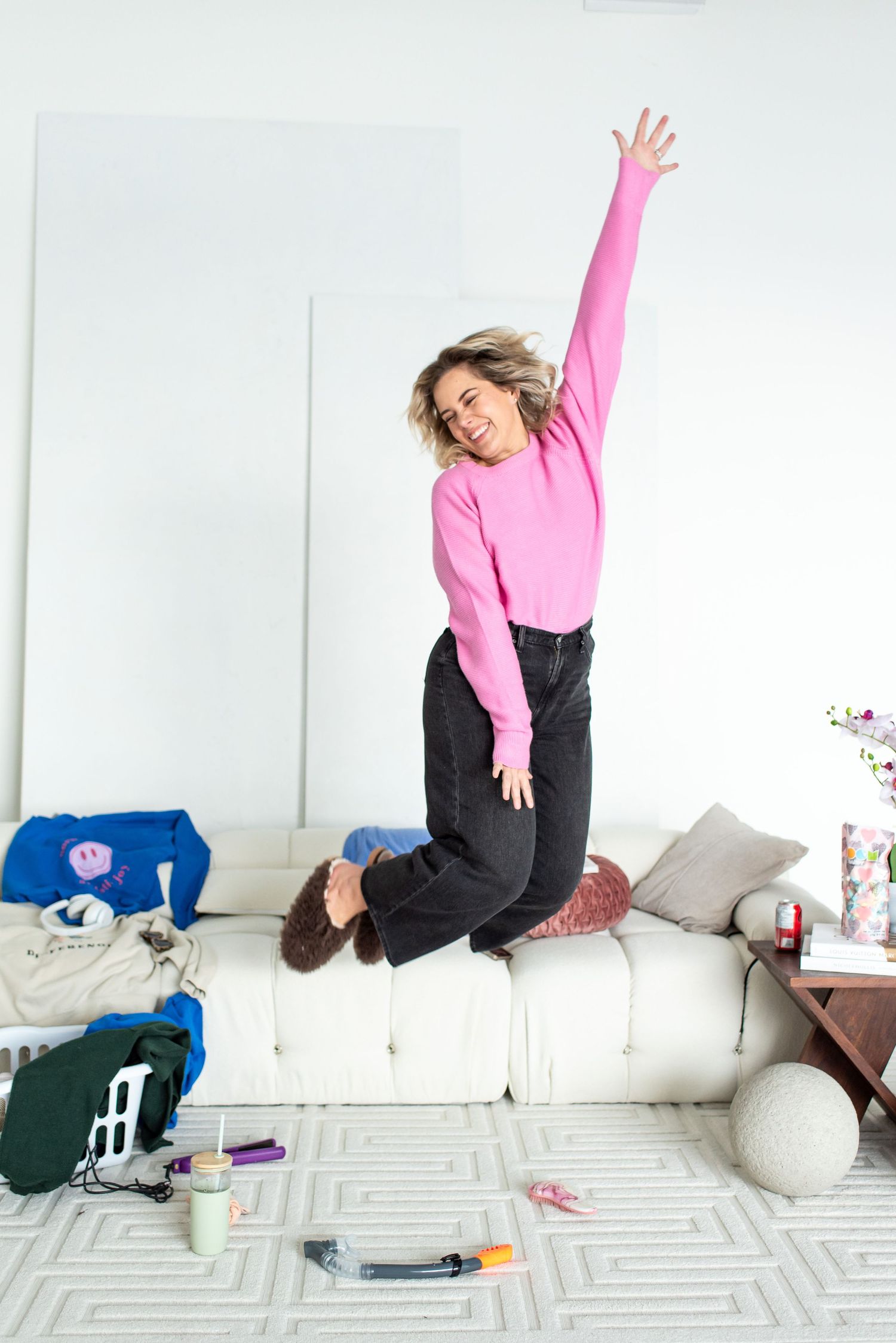 Energetic person in pink sweater and black pants jumping on white couch with scattered items on floor.
