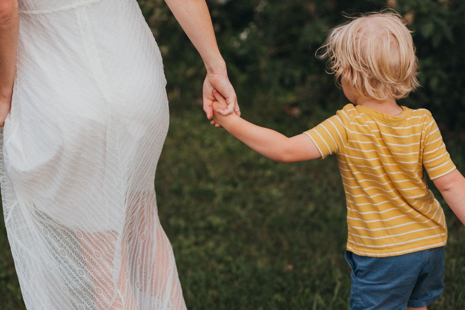 A child in a yellow striped shirt holds hands with someone wearing a flowing white dress in a garden setting.