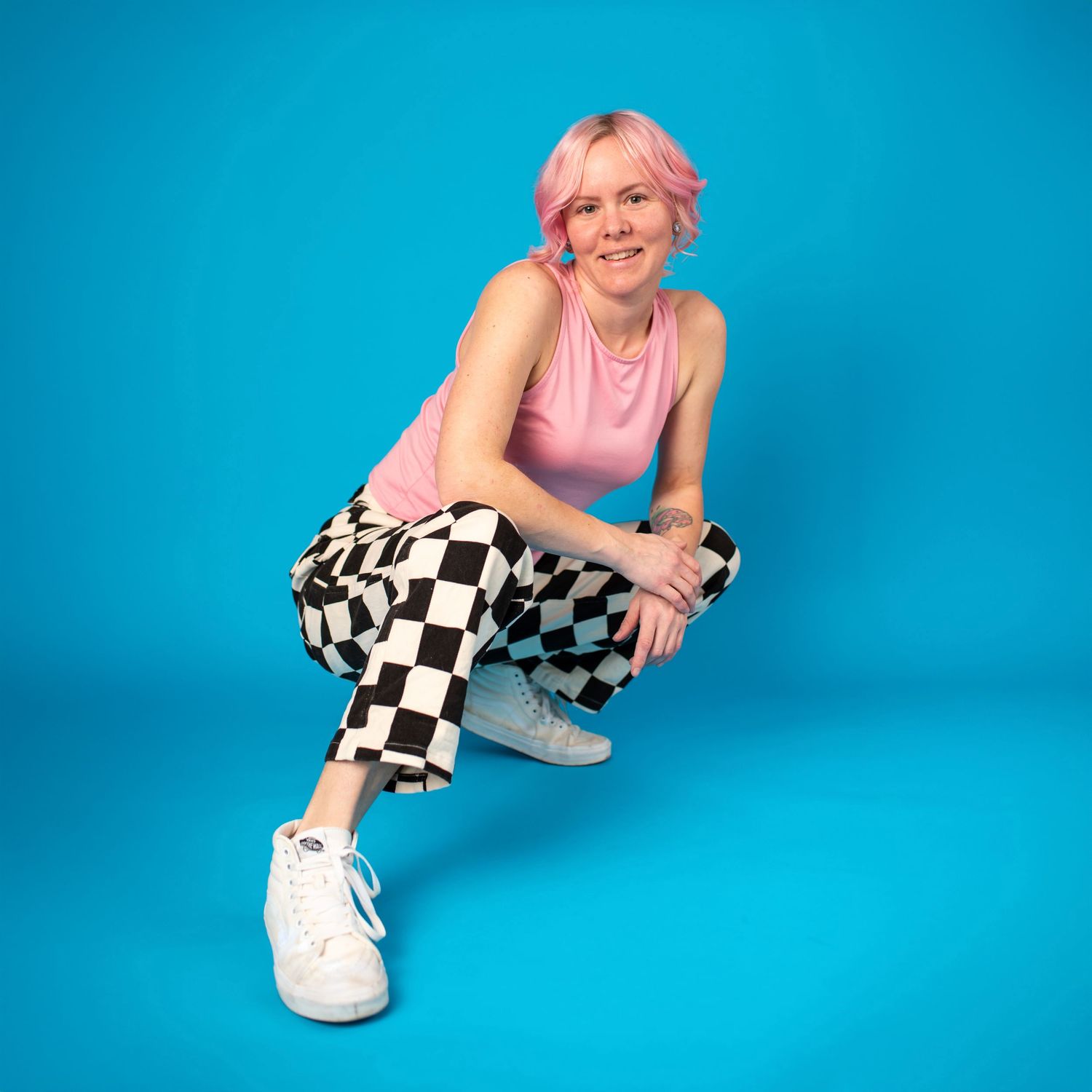 Person in pink sleeveless top and checkered pants posing against bright blue background, sitting with crossed legs.