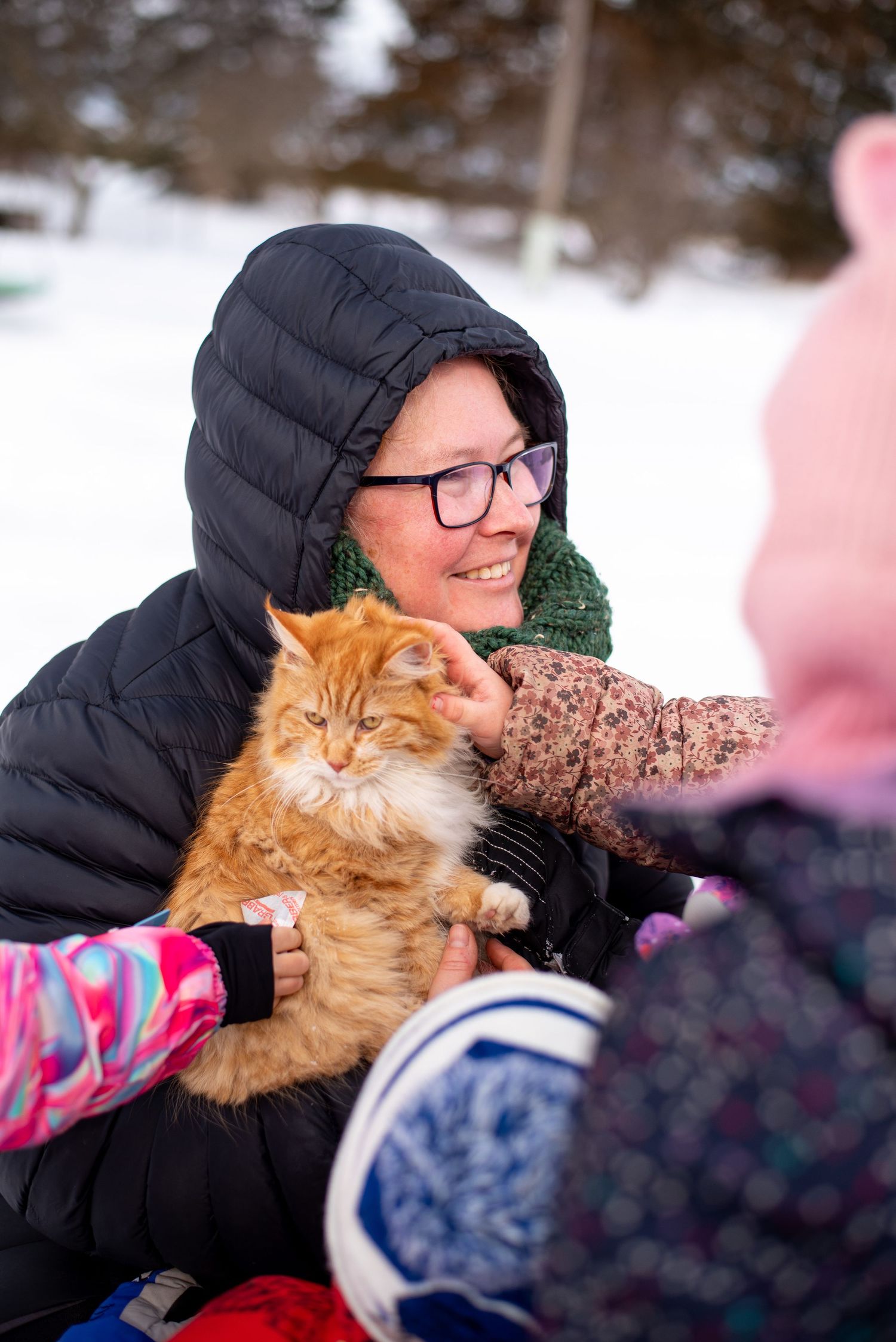 Someone in a black winter coat holds a fluffy orange cat while sitting outdoors in the snow.