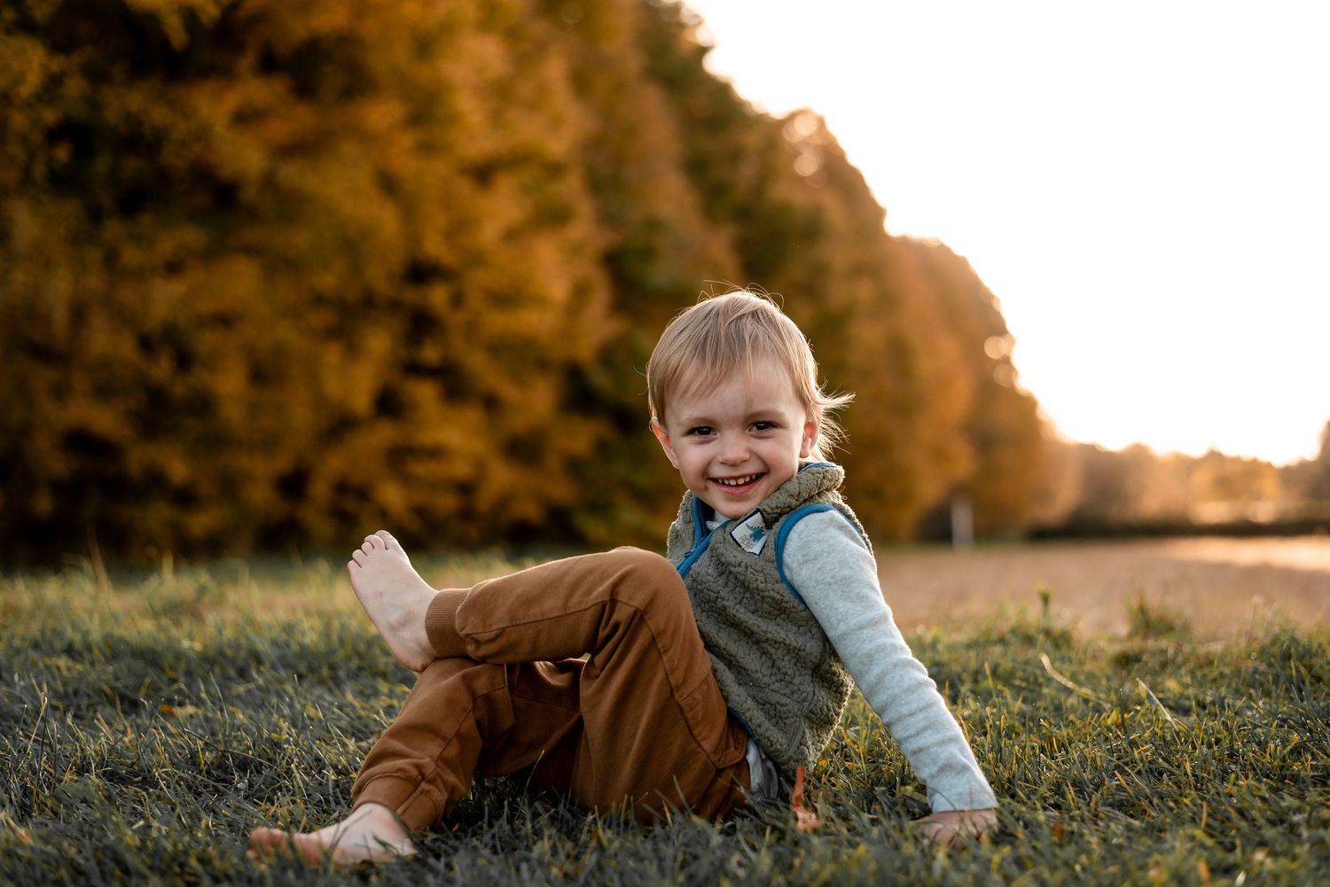 Child lying in golden grass field at sunset wearing light blue shirt and surrounded by autumn foliage.