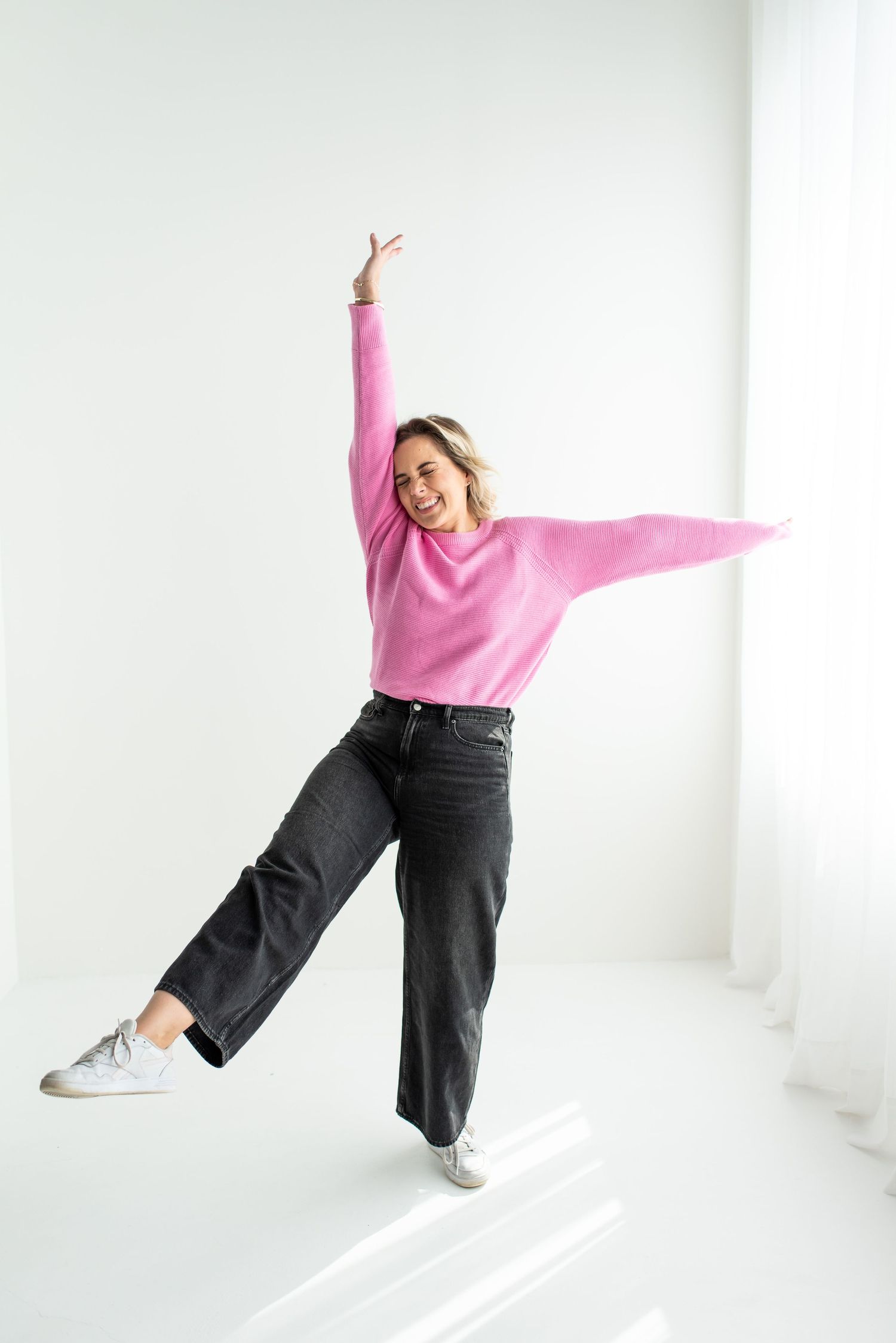 A joyful figure in a pink sweater and wide-leg jeans jumps playfully against a white background while striking a pose.
