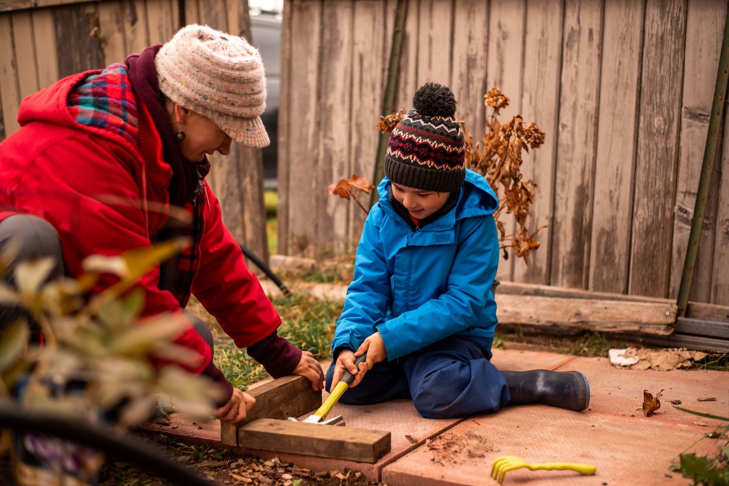 People in warm winter clothing work together on a gardening project near a wooden fence outdoors.