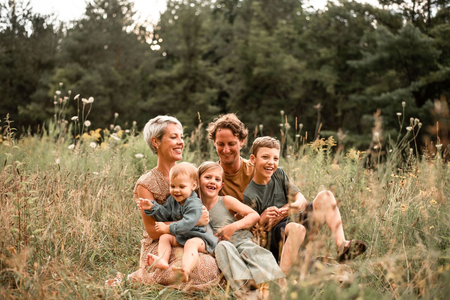 Candid family portrait in a sunny field with tall grass and pine trees.