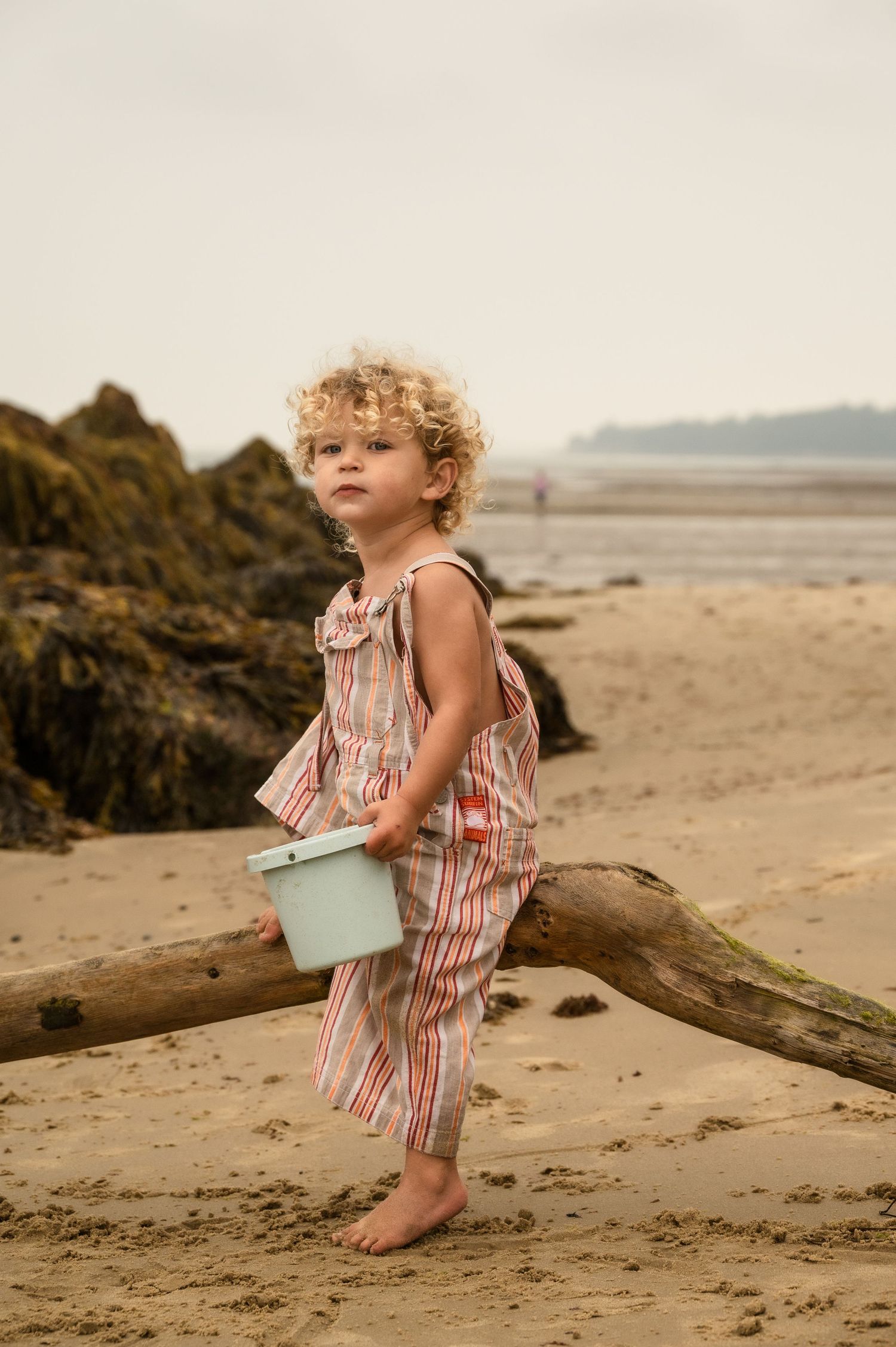 A young child in striped overalls carries a small blue bucket while exploring a rocky beach during sunset.