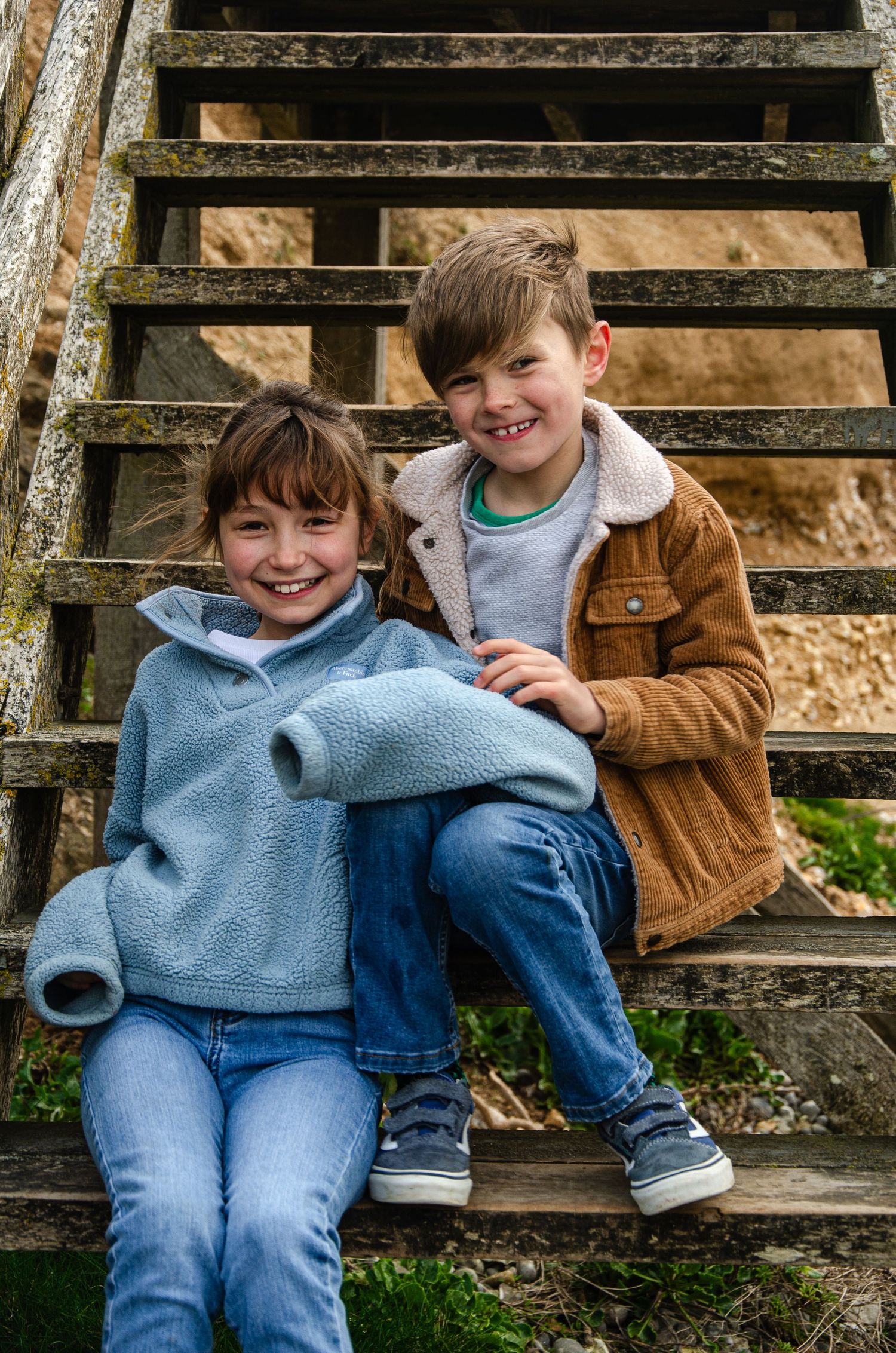 Two children sit smiling together on rustic wooden steps wearing casual clothing.