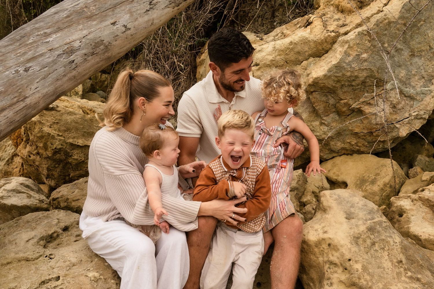 A family dressed in light colors sits together on rocky terrain while sharing a joyful moment outdoors.