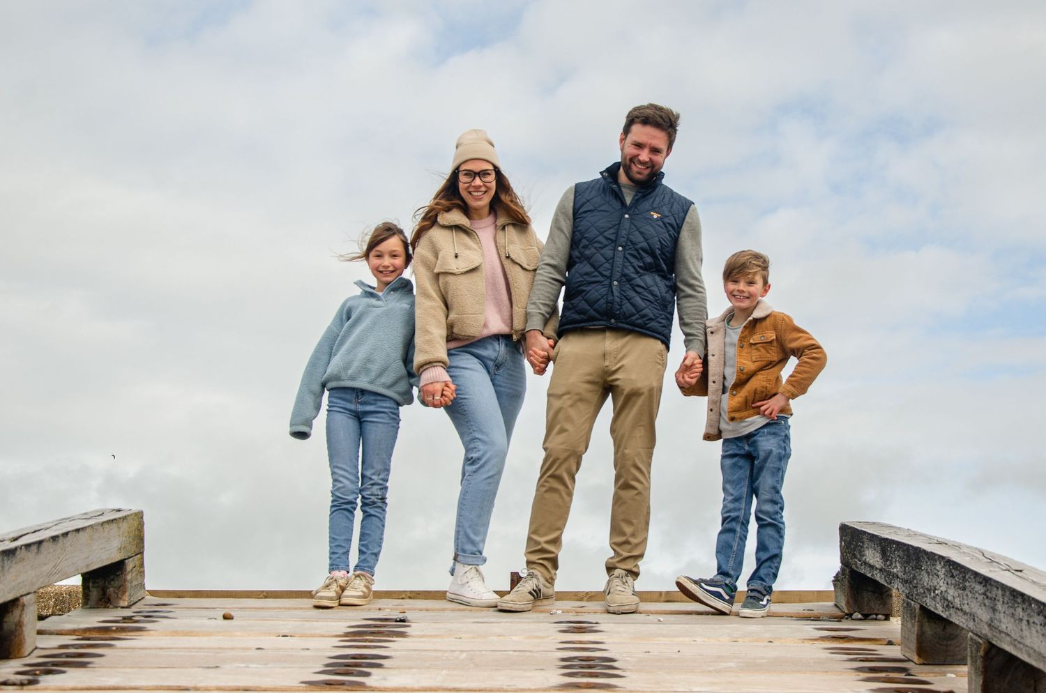 Family of four standing on wooden dock against cloudy sky.