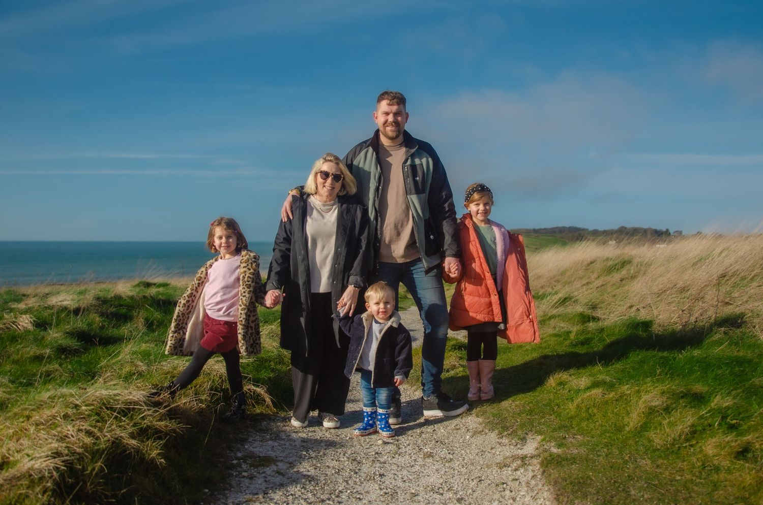 family with three young children standing holding hands on a grassy cliff and blue sky