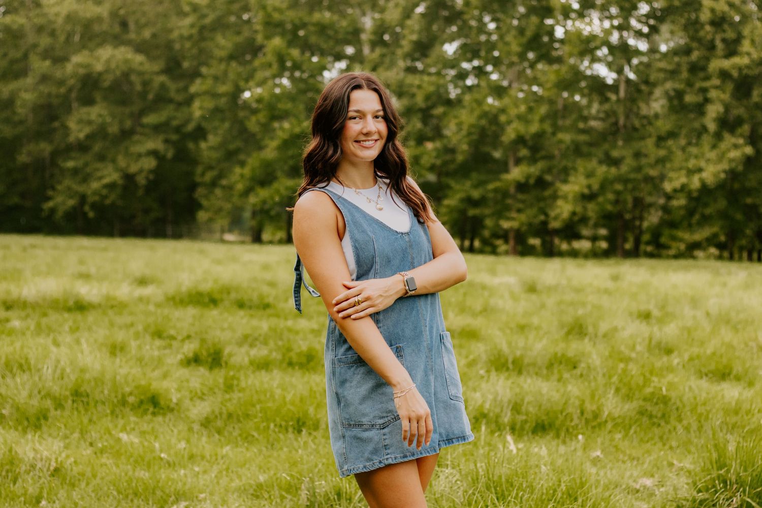 A senior in a blue denim dress smiles while standing in a sunny green field.