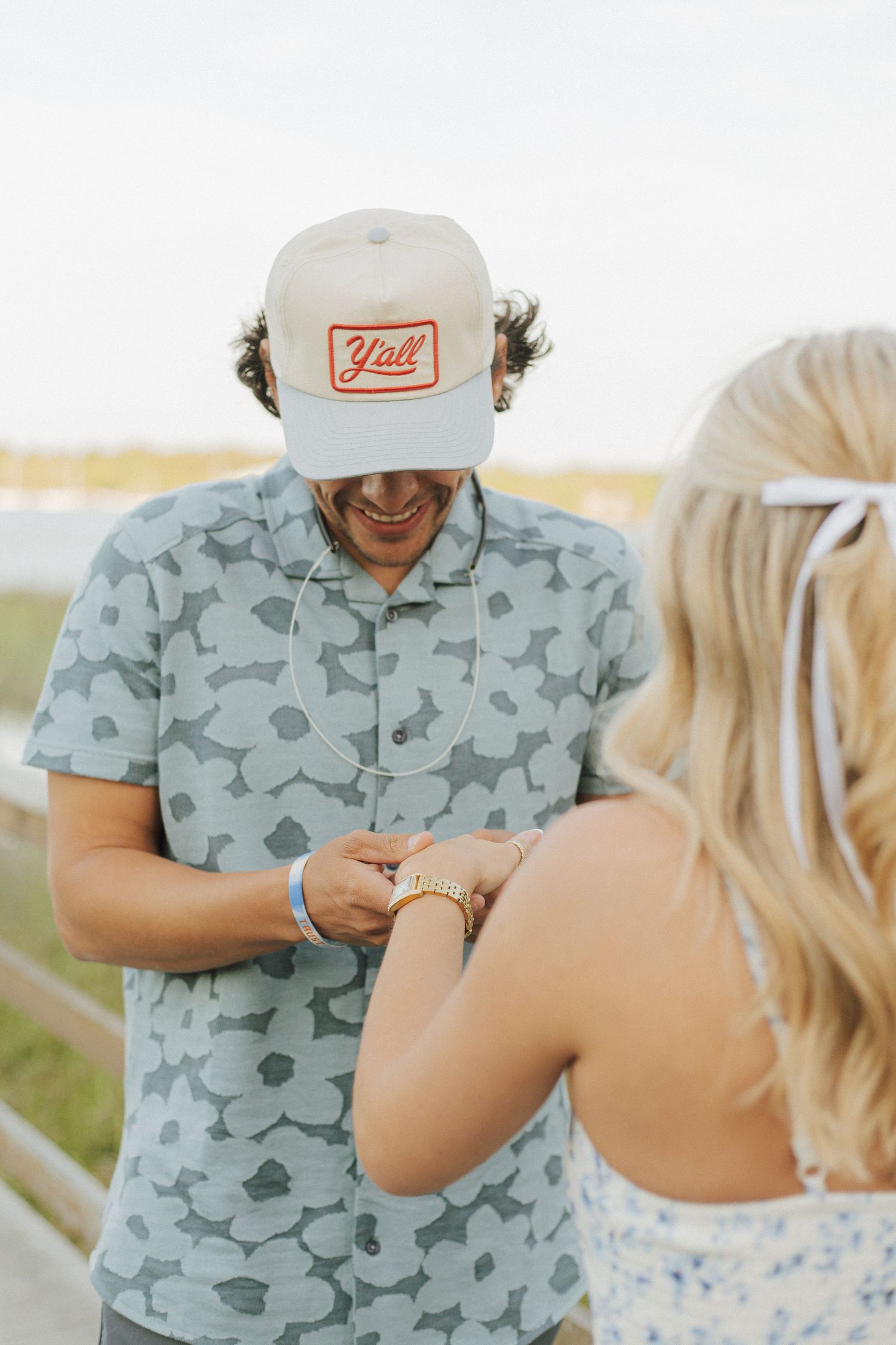 Man in a Y'all cap and floral shirt smiles while exchanging a gold ring with a blonde woman outdoors.