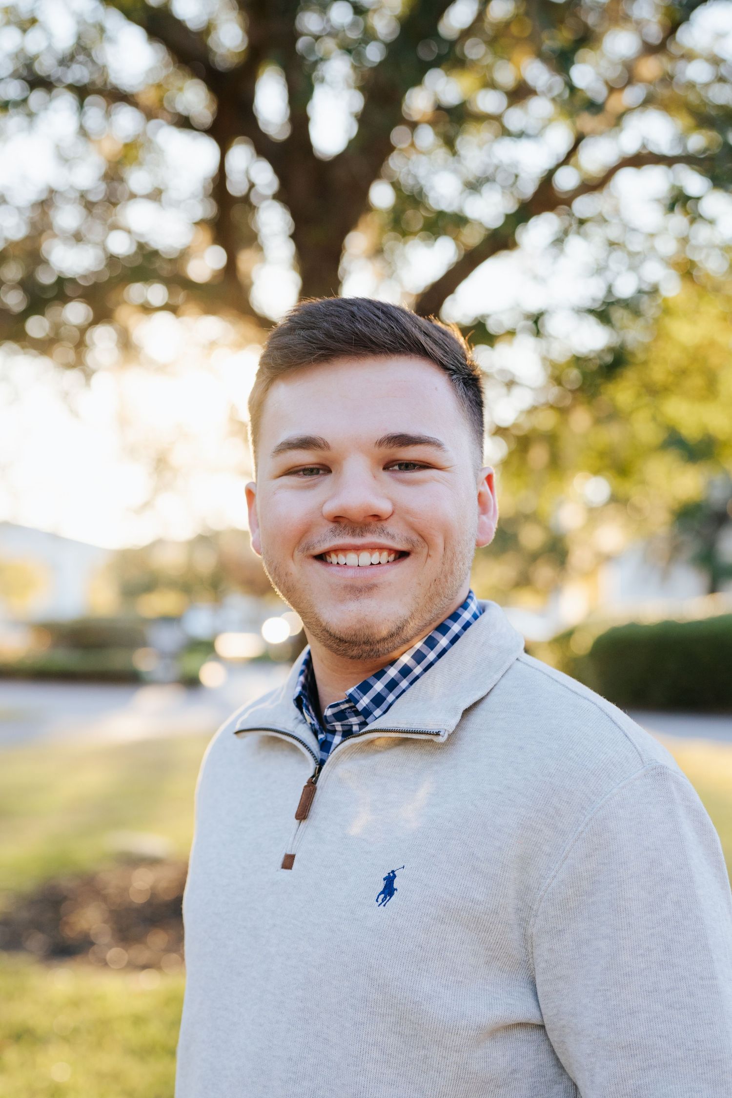 A person in a light gray quarter-zip pullover and checkered shirt poses outdoors with trees and sunlight in the background.