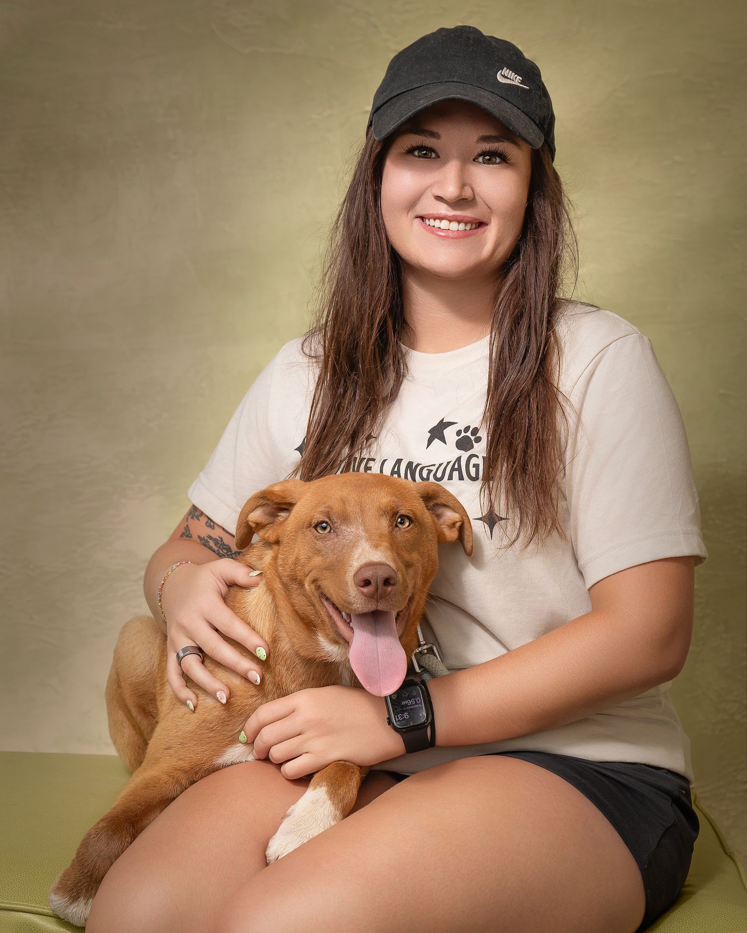 Young woman in Nike cap and paw print tee smiling while hugging a happy brown dog with tongue out.